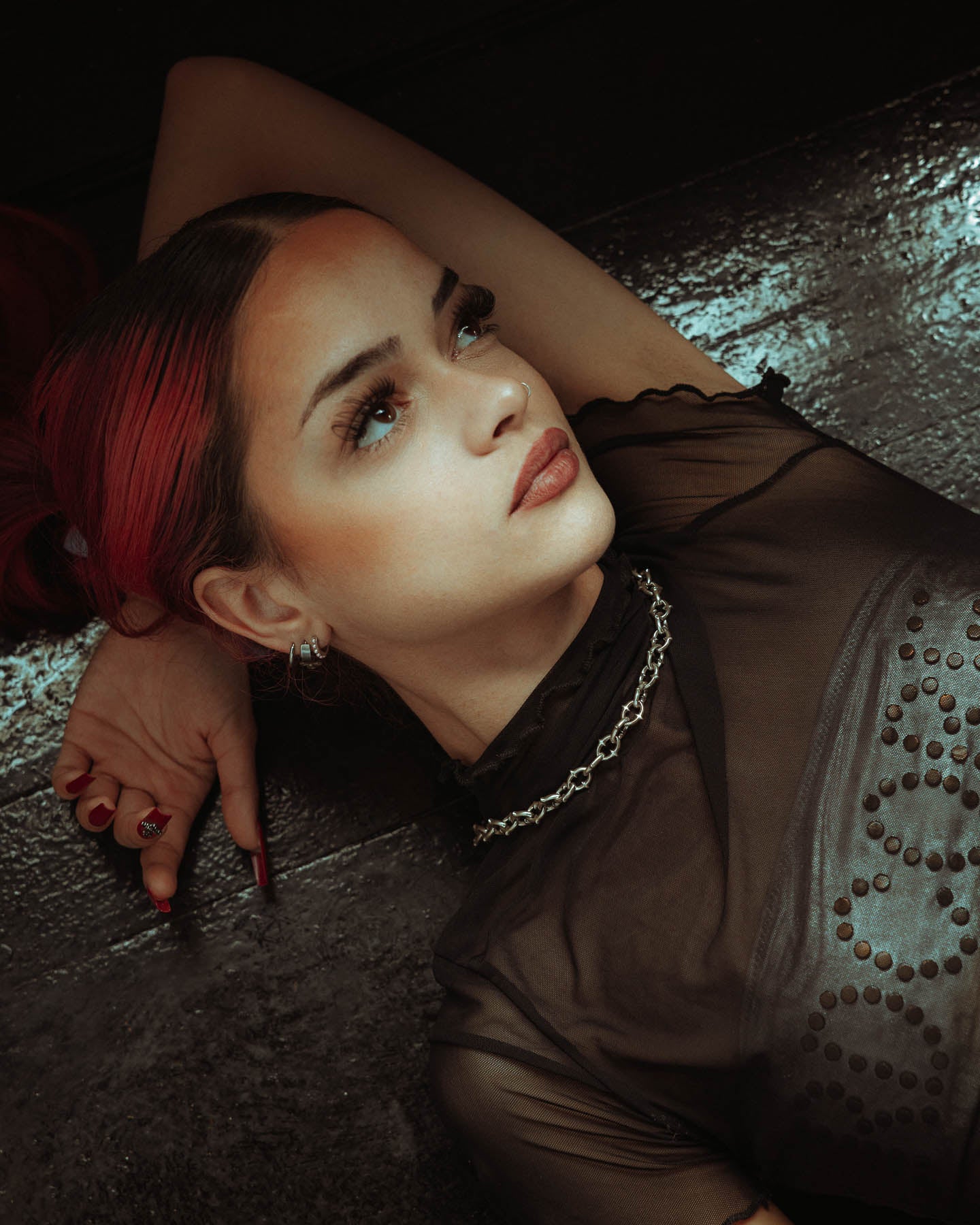 A young woman with red-tinted hair lies on a dark surface, resting her head on her arm. She gazes thoughtfully, wearing a sheer black dotted top and the 925 Sterling Silver "The Cathedral" 9mm Spiked Chain.