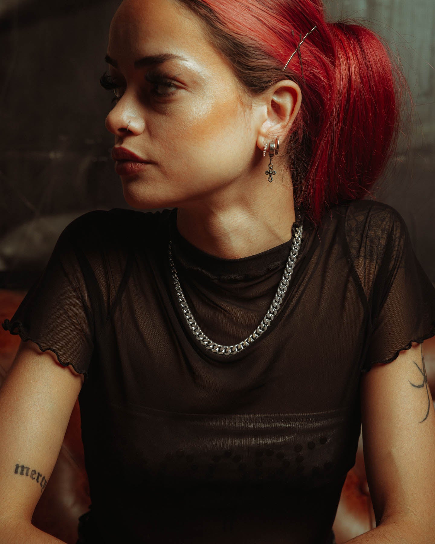A red-haired woman with her hair tied back wears a sheer black top and a 925 Sterling Silver 9mm Cuban Chain necklace. She has hoop and cross earrings, a nose ring, tattoos on her arms, and sits on a brown seat looking left.