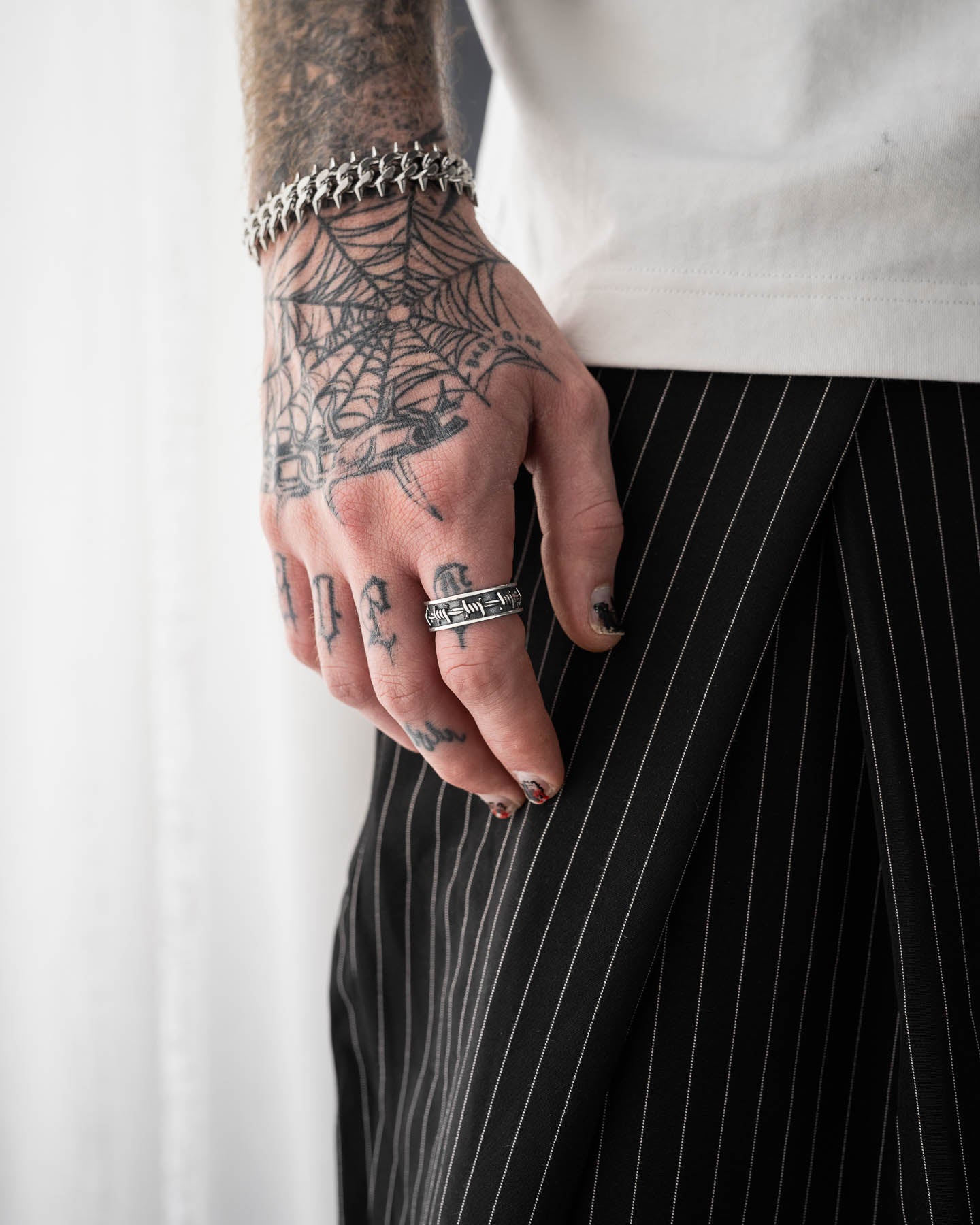 A tattooed hand with stackable rings and the 925 Sterling Silver Barbed Wire Ring rests beside a person in a white shirt and black pinstripe pants, featuring bold accessories, dark nail polish, and finger tattoos.
