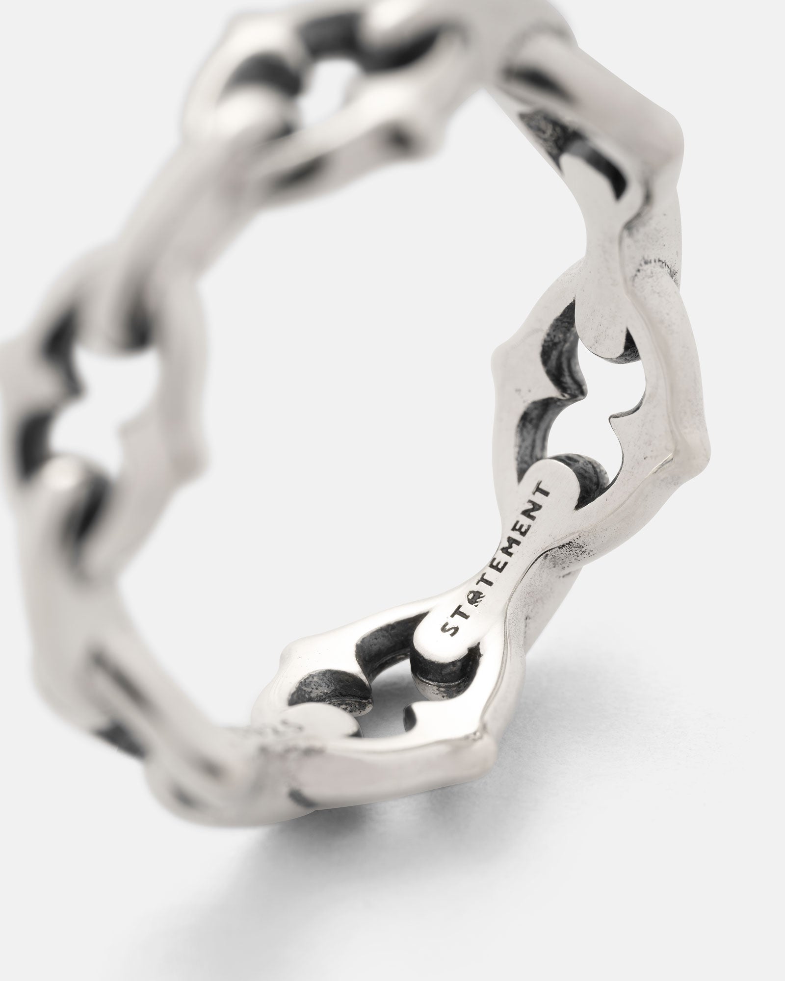A close-up of the 925 Sterling Silver "The Cathedral" Spiked Ring shows its geometric chain link design. The inner band is engraved with the word STATEMENT, highlighting the texture and intricate details.