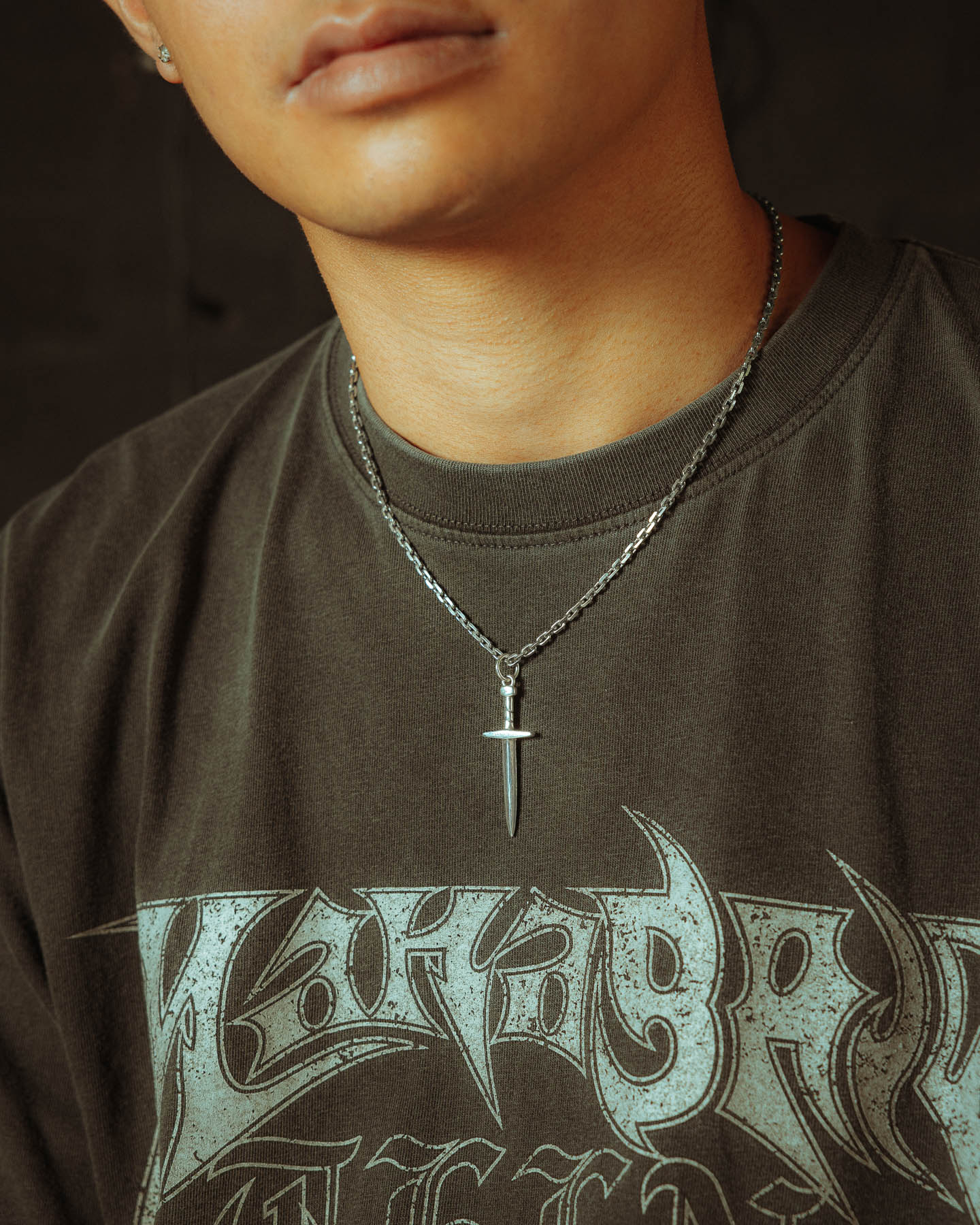 A person wearing a dark graphic T-shirt and a silver chain with the 925 Sterling Silver Dagger Pendant; the image is cropped to show only the lower face, neck, and upper chest.
