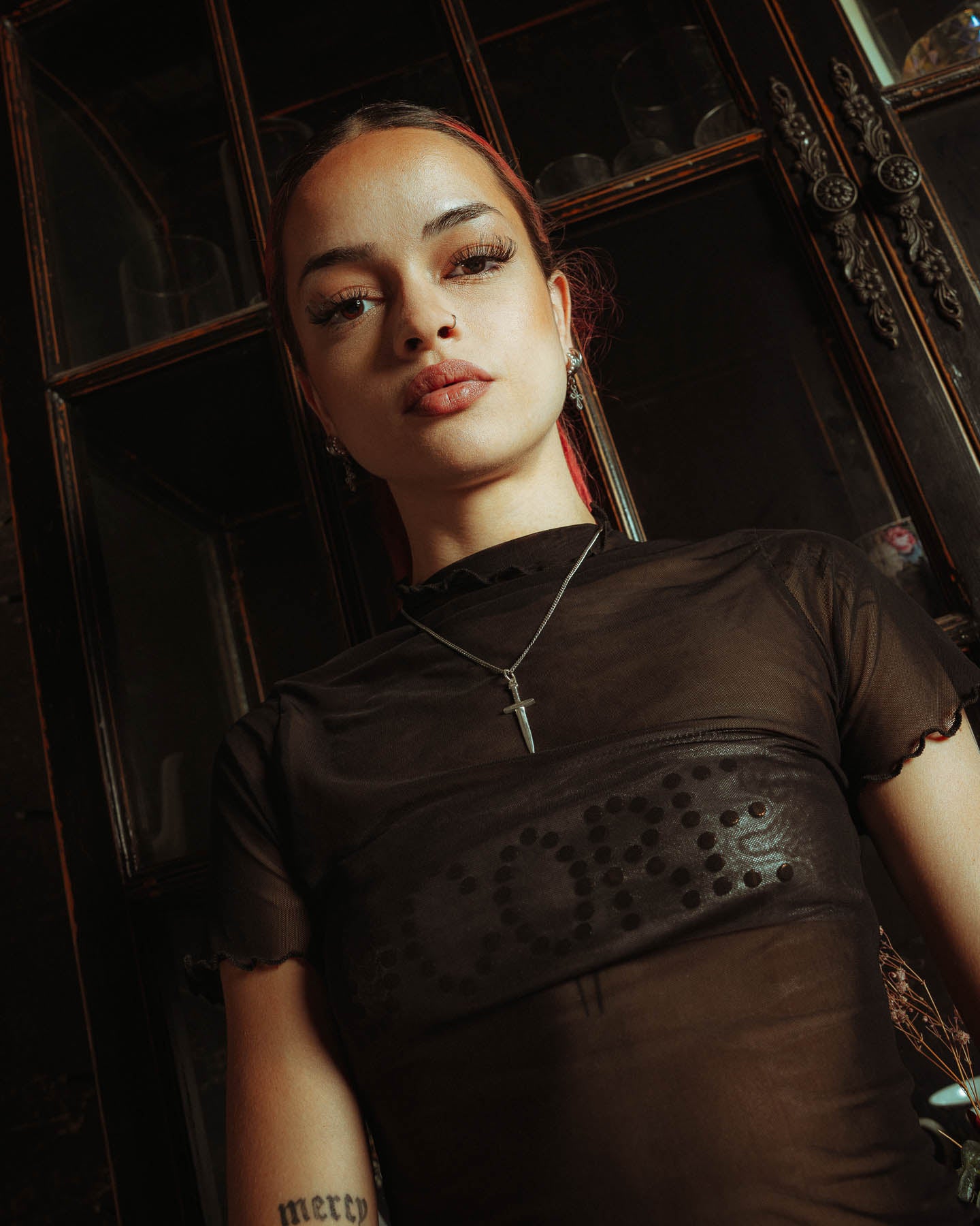 A woman with long eyelashes and a neutral expression stands before a glass cabinet, wearing a sheer black top and the 925 Sterling Silver Dagger Pendant. The photo is shot from a low angle.