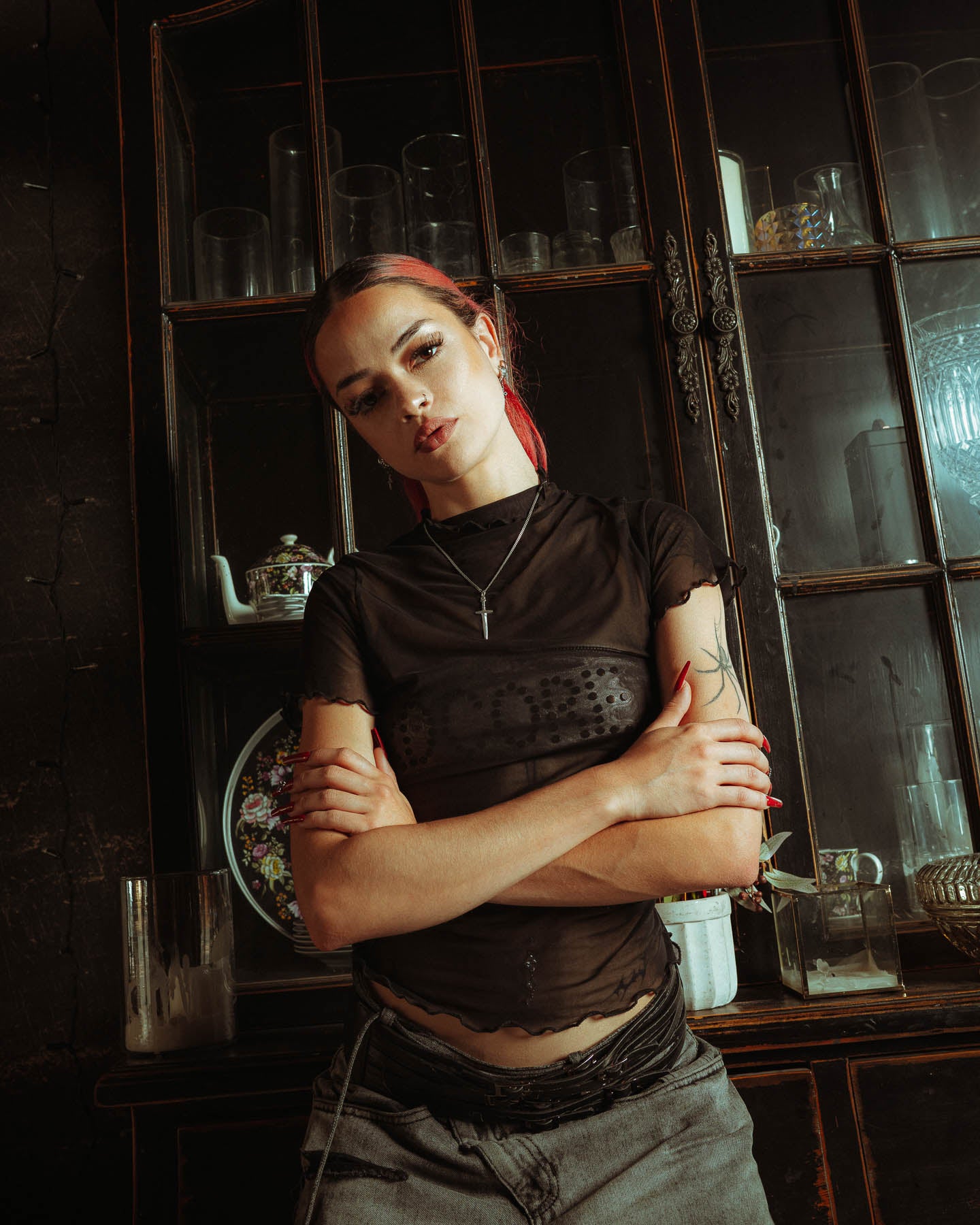 A red-haired person, in a sheer black top and gray pants, stands with arms crossed wearing a handmade 925 Sterling Silver Dagger Pendant, in front of a vintage glass cabinet filled with glassware, teapots, and decorative plates.