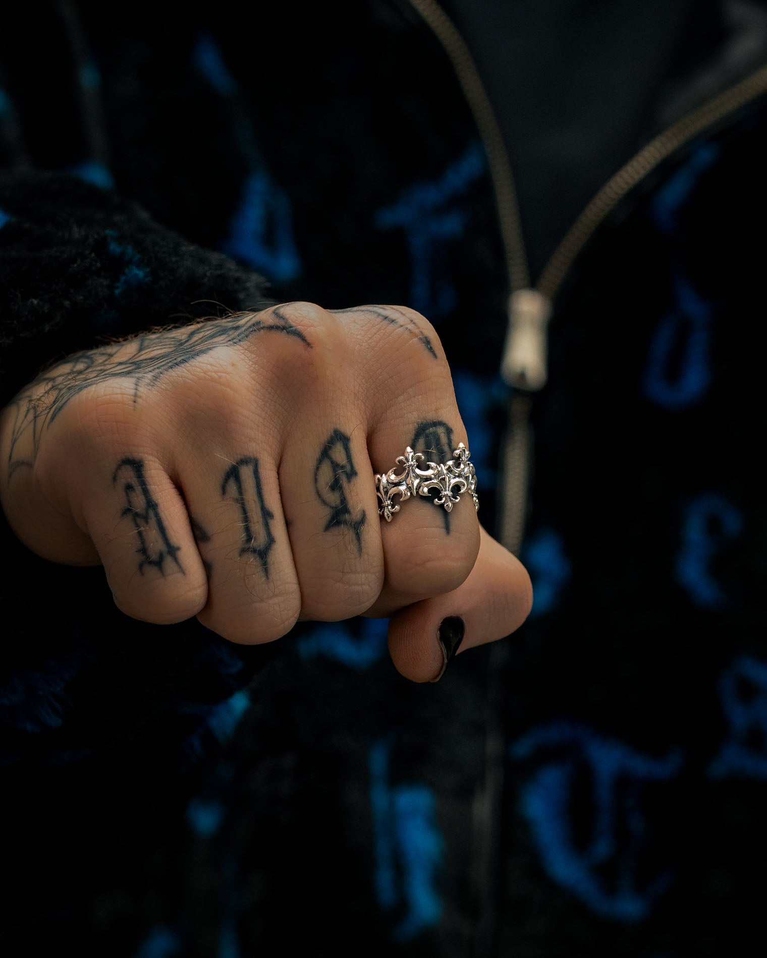 A close-up of a tattooed hand with ALIVE inked on the fingers, adorned with the Statement Collective 925 Sterling Silver “Crested” Fleur De Lis Ring, featuring an ornate vintage design. The person is dressed in a dark, textured jacket with a blue pattern.