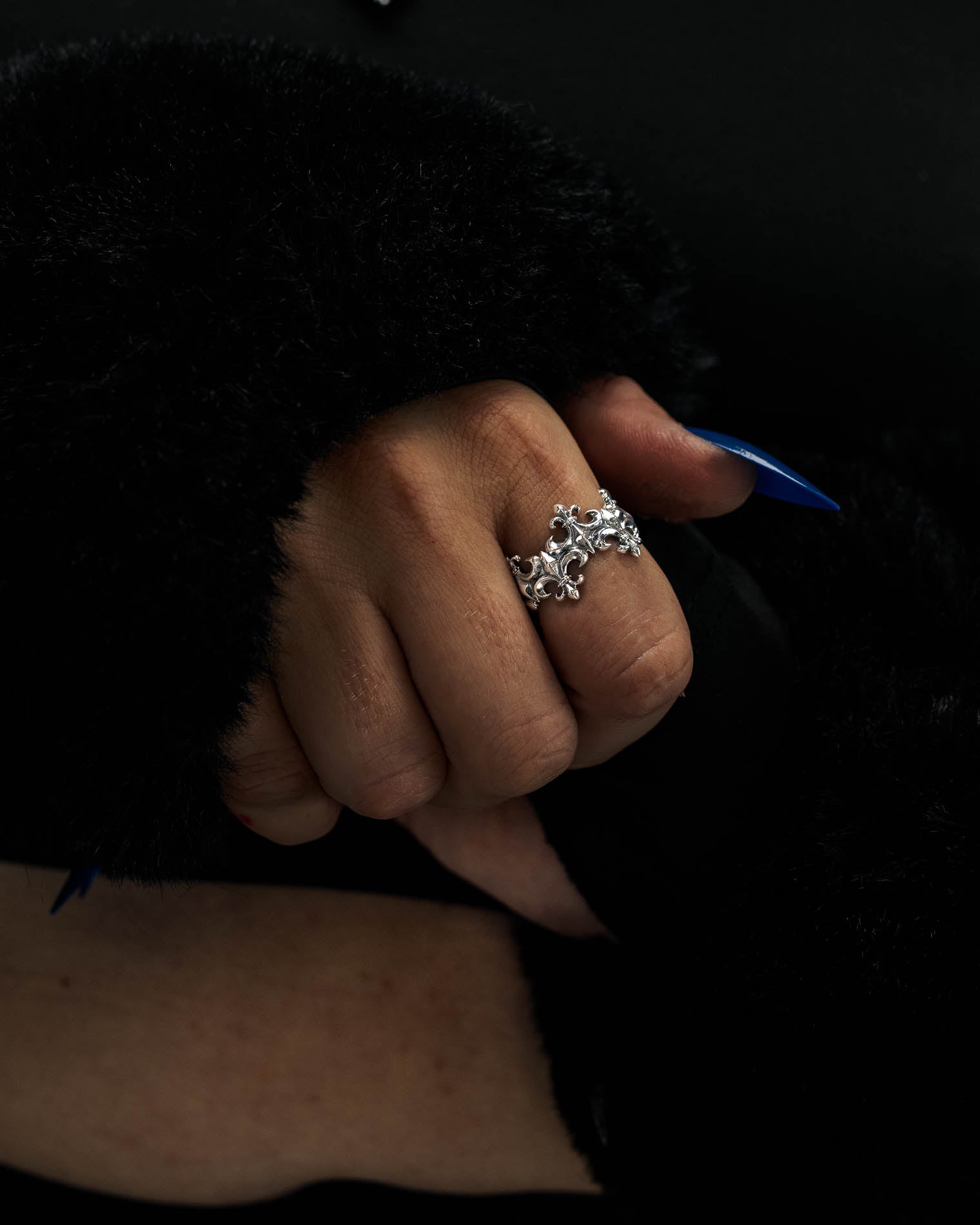 A hand with blue nails, adorned by the Statement Collective 925 Sterling Silver “Crested” Fleur De Lis Ring, is shown against a dark, fuzzy sleeve. The ring’s ornate design with round and marquise-shaped stones enhances its allure as vintage-inspired silver jewelry.
