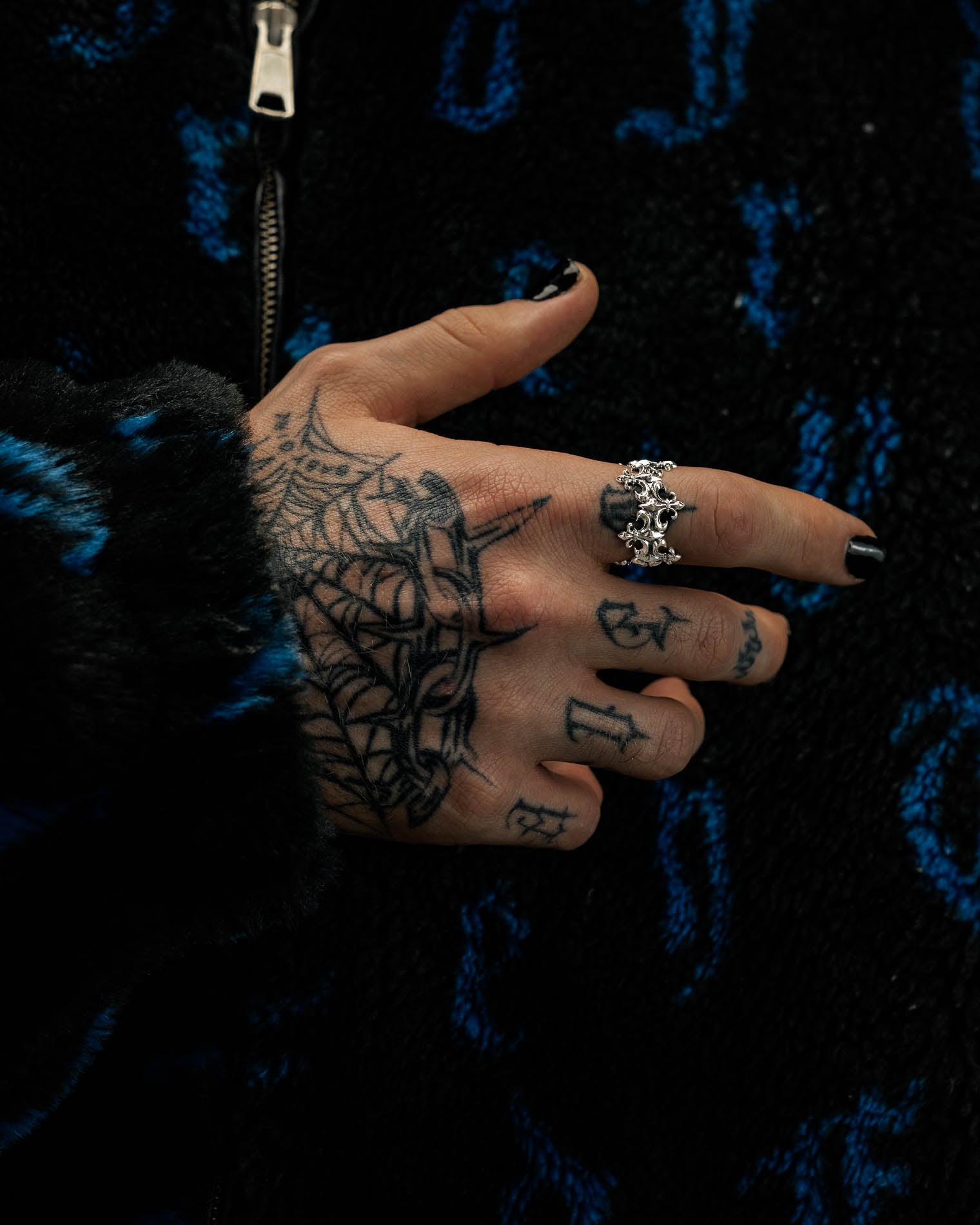 A hand with black nail polish, a spider web tattoo, and various small tattoos wears the 925 Sterling Silver “Crested” Fleur De Lis Ring by Statement Collective. The person is wearing a black and blue fuzzy jacket with a zipper.