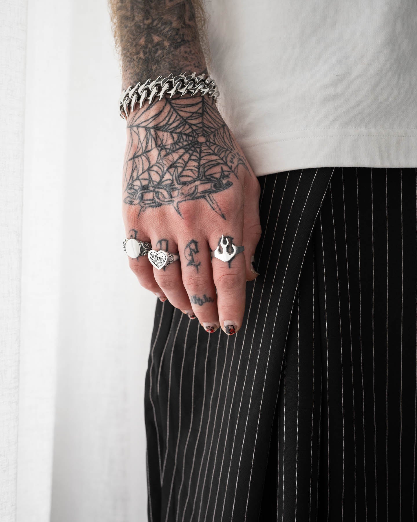 A tattooed hand with a spiderweb design and various rings, including the 925 Sterling Silver Flame Ring, rests against a pinstriped black skirt. The person wears a chunky chain bracelet, white top, and has black nails tipped in red.