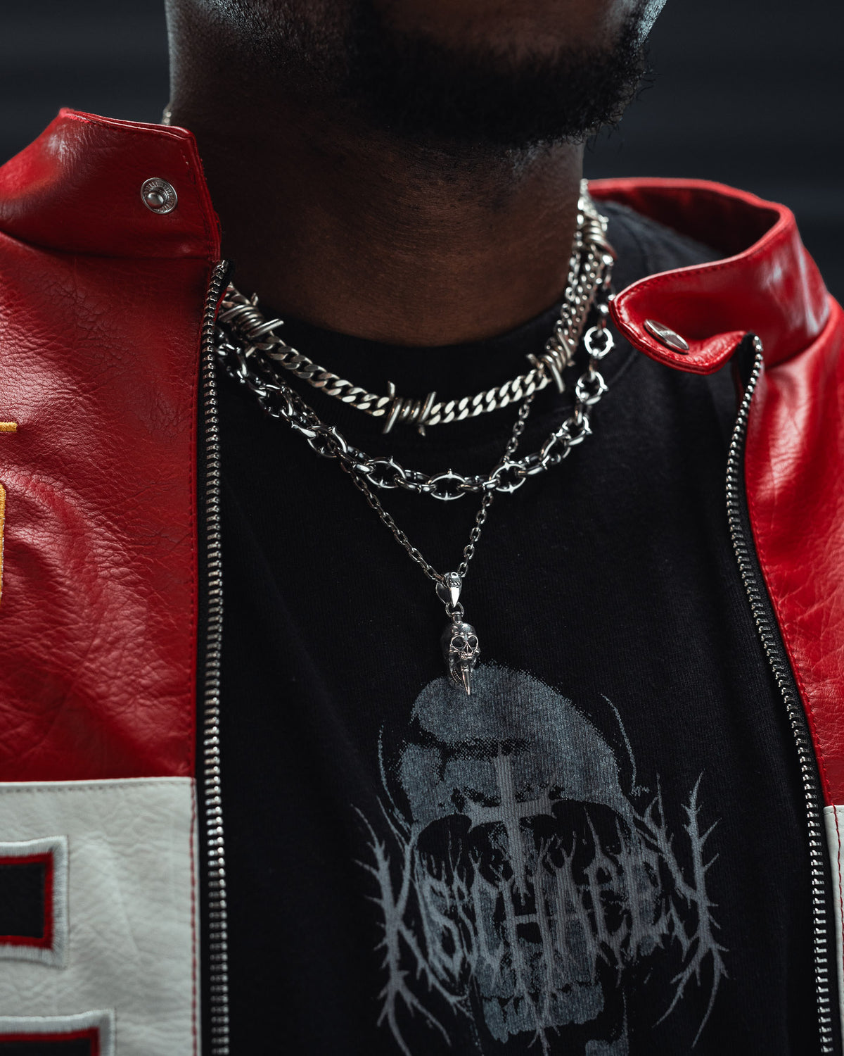 Close-up of a person wearing layered silver chains, including the Statement Collective 925 Sterling Silver Mini “Bite the Bullet” Pendant necklace, a red leather jacket with a white patch, and a black shirt featuring a skull and gothic-style lettering. The person’s face is not visible.