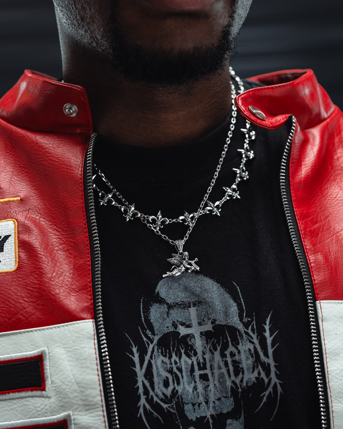 A person is shown from the shoulders to the neck wearing a red and white jacket, a black t-shirt with gothic artwork, and two silver chain necklaces—one adorned with cross-shaped pendants and the other featuring Statement Collective’s 925 Sterling Silver Mini “Deaths Cupid” Pendant.