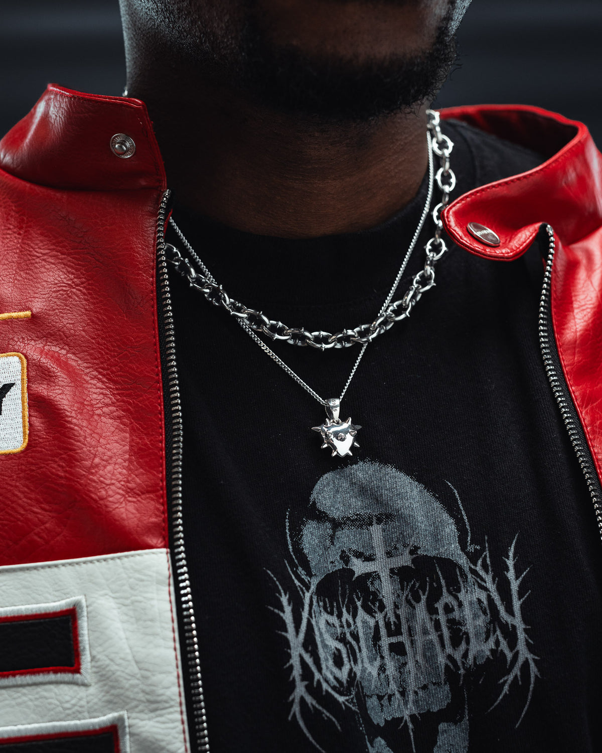 A person wearing a red leather jacket, layered silver necklaces—including the striking Statement Collective 925 Sterling Silver Mini “Love Hurts” Pendant—and a black graphic T-shirt with a skull and gothic text. This stylish ensemble channels dark romance jewelry vibes. The photo is cropped to show only the torso and neck.