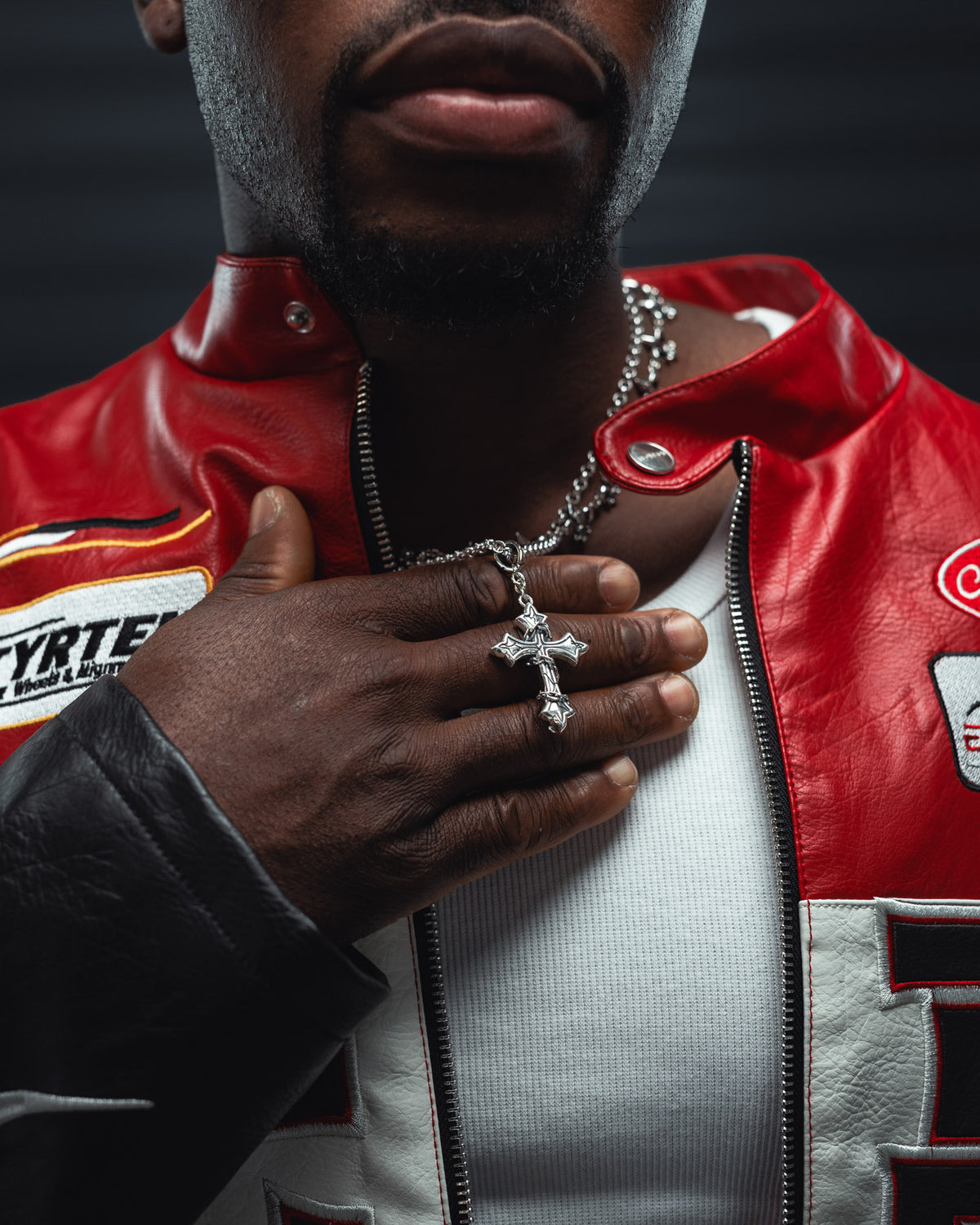 A person wearing a red and white jacket adorned with patches holds the Statement Collective 925 Sterling Silver Mini “The Ruins” Cross Pendant against their chest. A white shirt peeks out from beneath the jacket, while only their lower face and hand are visible—emphasizing the elegant gothic pendant as it gleams in their grip.