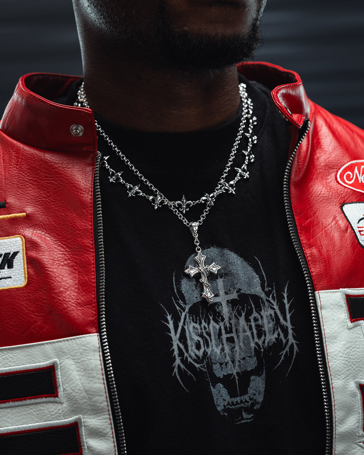 A person wears a red and white jacket, a black shirt with a skull graphic, and layered silver necklaces—including Statement Collective’s 925 Sterling Silver Mini “The Ruins” Cross Pendant. The wrapped-chain design of the necklace lends an edgy, subtle gothic vibe to the look. The image is cropped to focus on the lower face, neck, and chest.