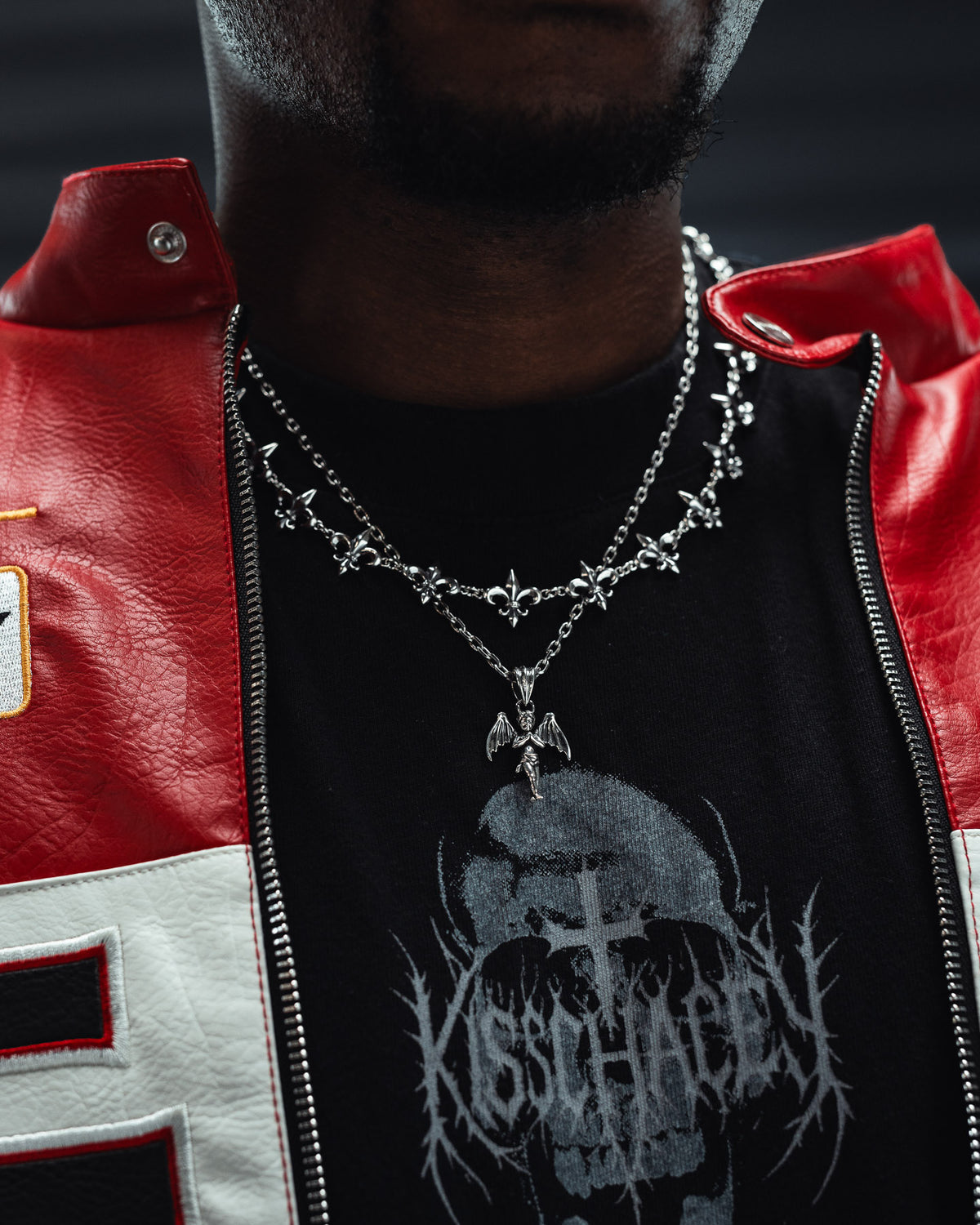 A person in a red and white jacket wears layered sterling silver necklaces, including the Statement Collective 925 Sterling Silver “The Wicked One” Pendant, paired with a black shirt featuring a gothic skull and text design. The focus is on the punk jewelry and bold shirt.