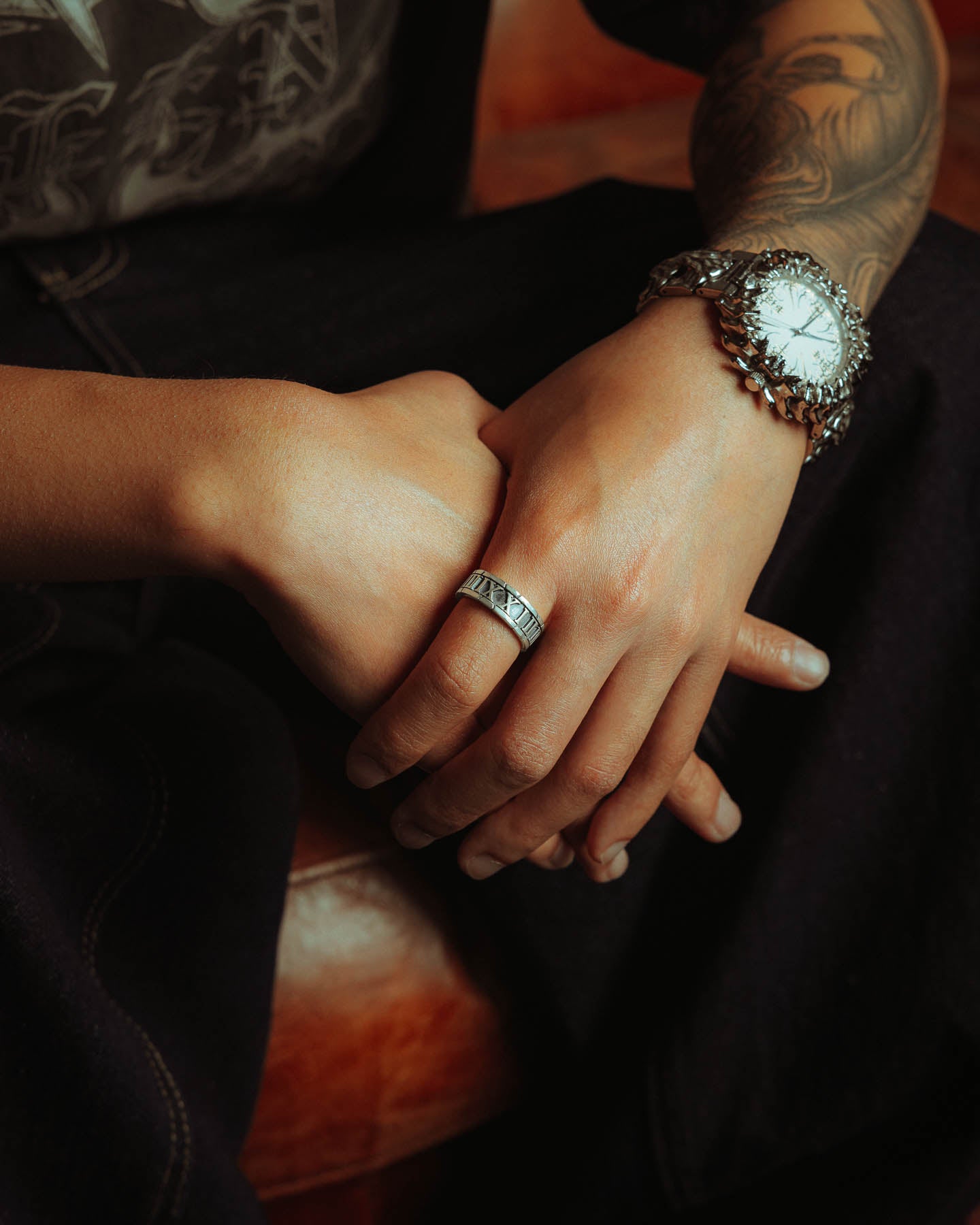 A person with a tattooed arm wears a large wristwatch and the 925 Sterling Silver Numerals Ring, resting their hands on their lap against dark clothing and a brown leather surface.