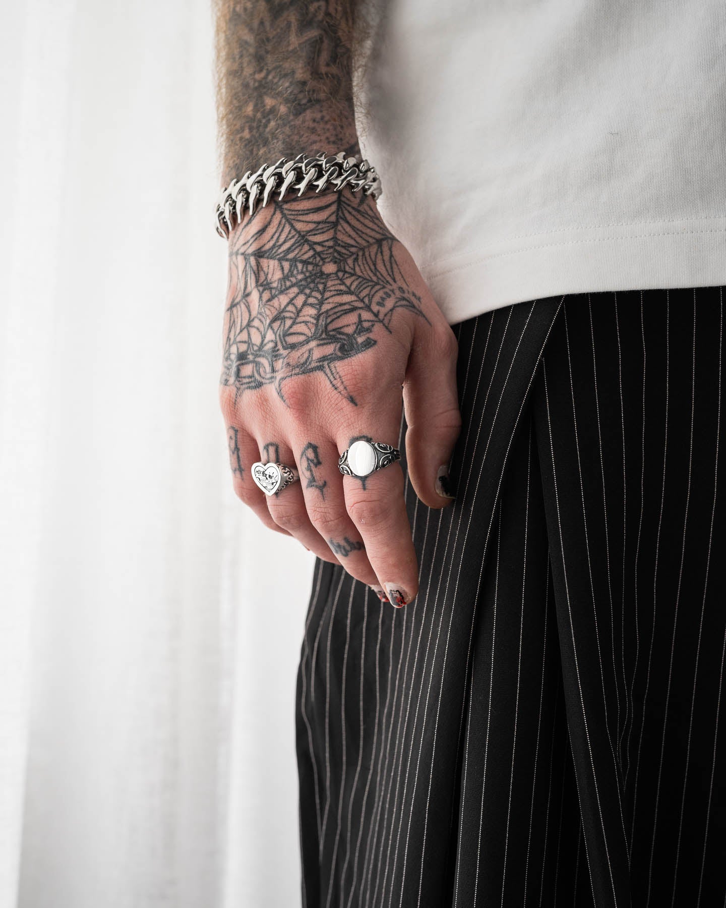 A tattooed hand adorned with the 925 Sterling Silver Mini Oval Scroll Signet Ring and a chunky chain bracelet rests by their side. The person wears a white shirt and black pinstripe pants, set against softly lit white curtains.