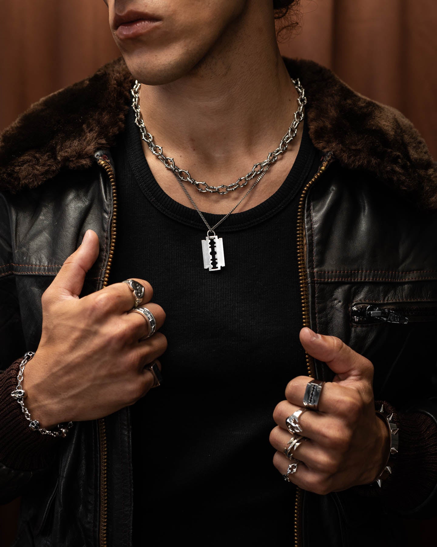 Against a brown background, a person wears a black shirt, dark leather jacket with fur collar, punk metal jewelry, and layered chain necklaces—including the 925 Sterling Silver Razor Blade Pendant.