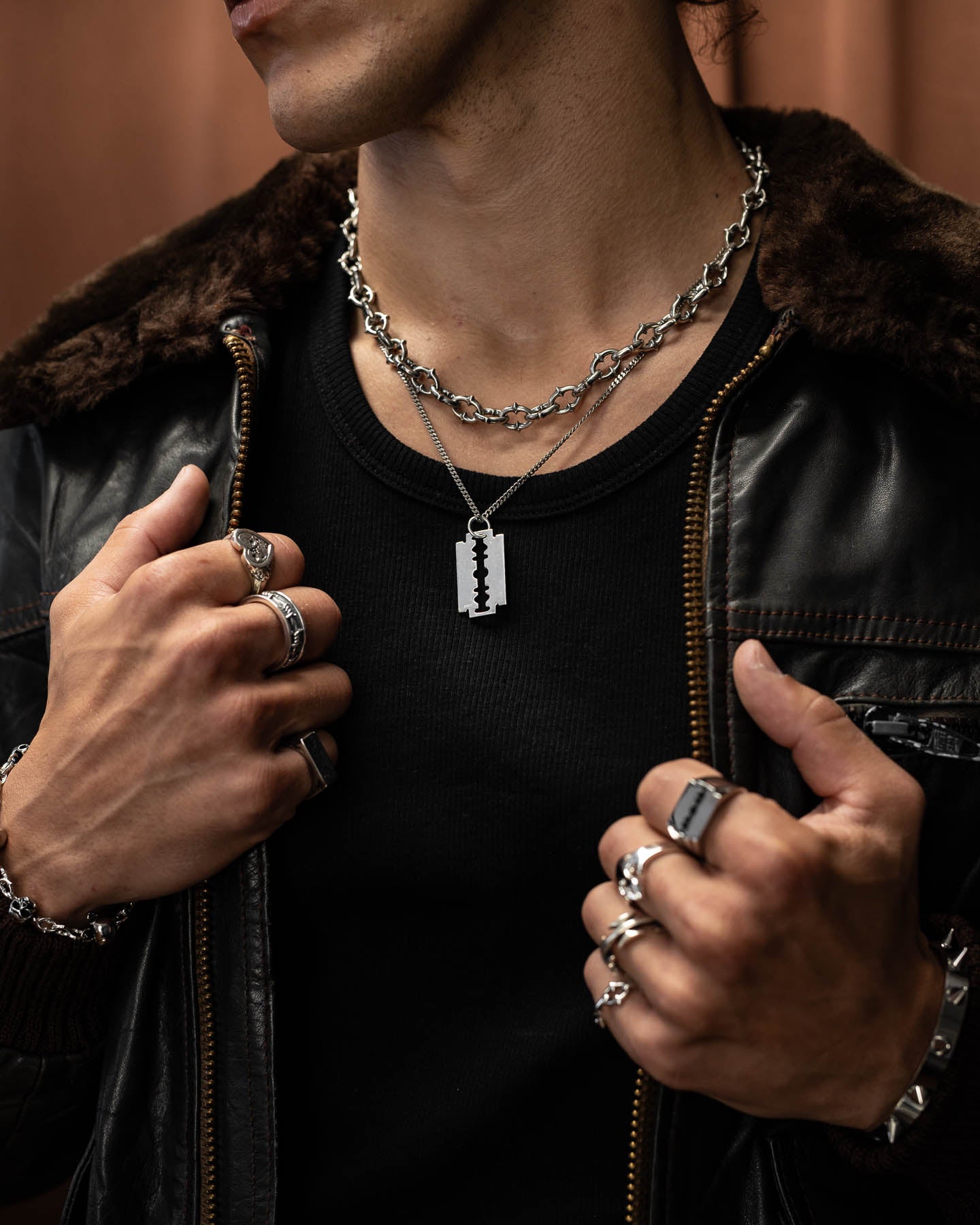 A person wearing a black shirt and leather jacket with fur collar, accessorized with punk metal jewelry—multiple rings, chain bracelets, and layered necklaces, including the 925 Sterling Silver Razor Blade Pendant.