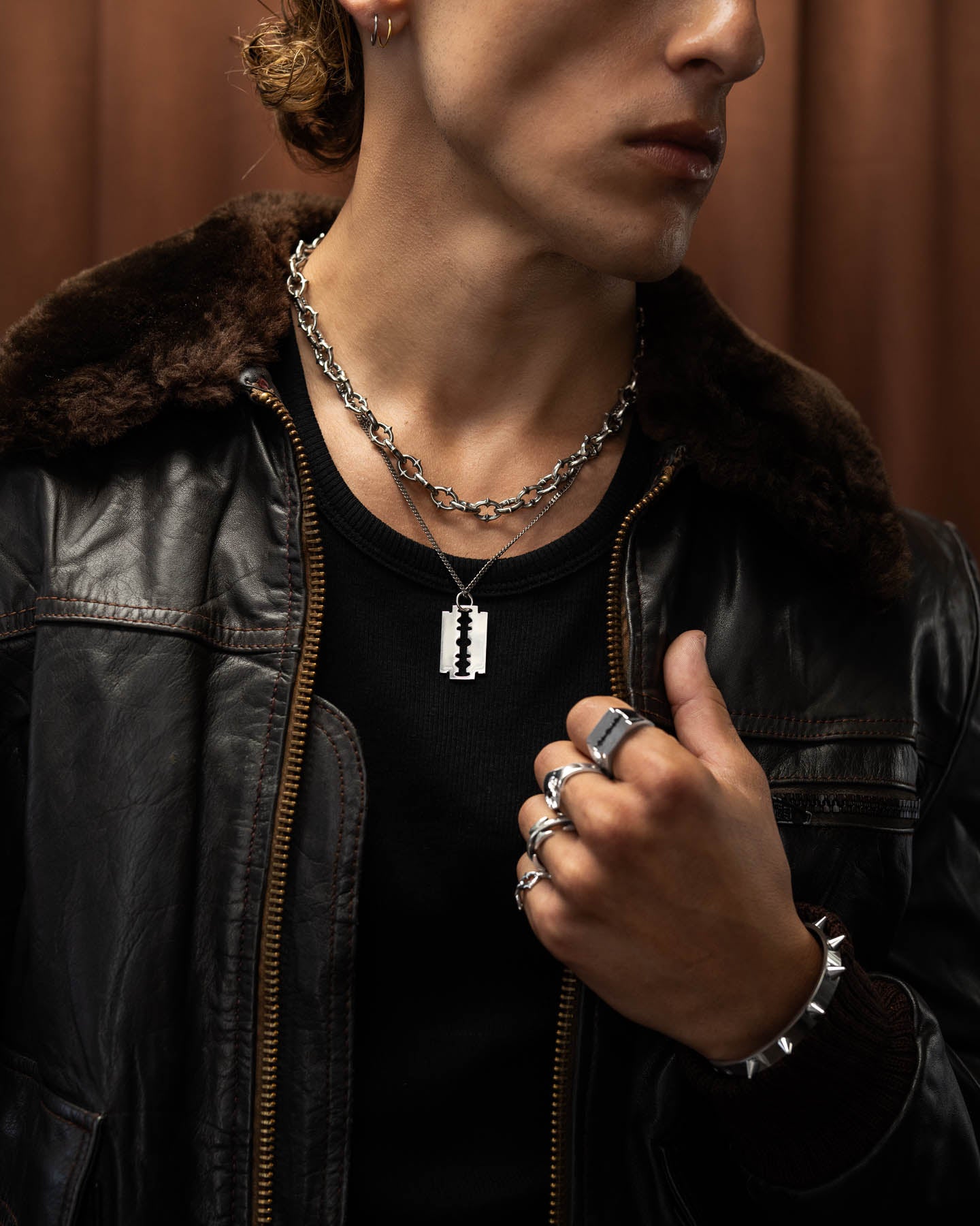 A person wearing a black leather jacket with a fur collar, layered punk metal jewelry including the 925 Sterling Silver Razor Blade Pendant, along with chunky metal rings and bracelets, holds their hand near their chest.