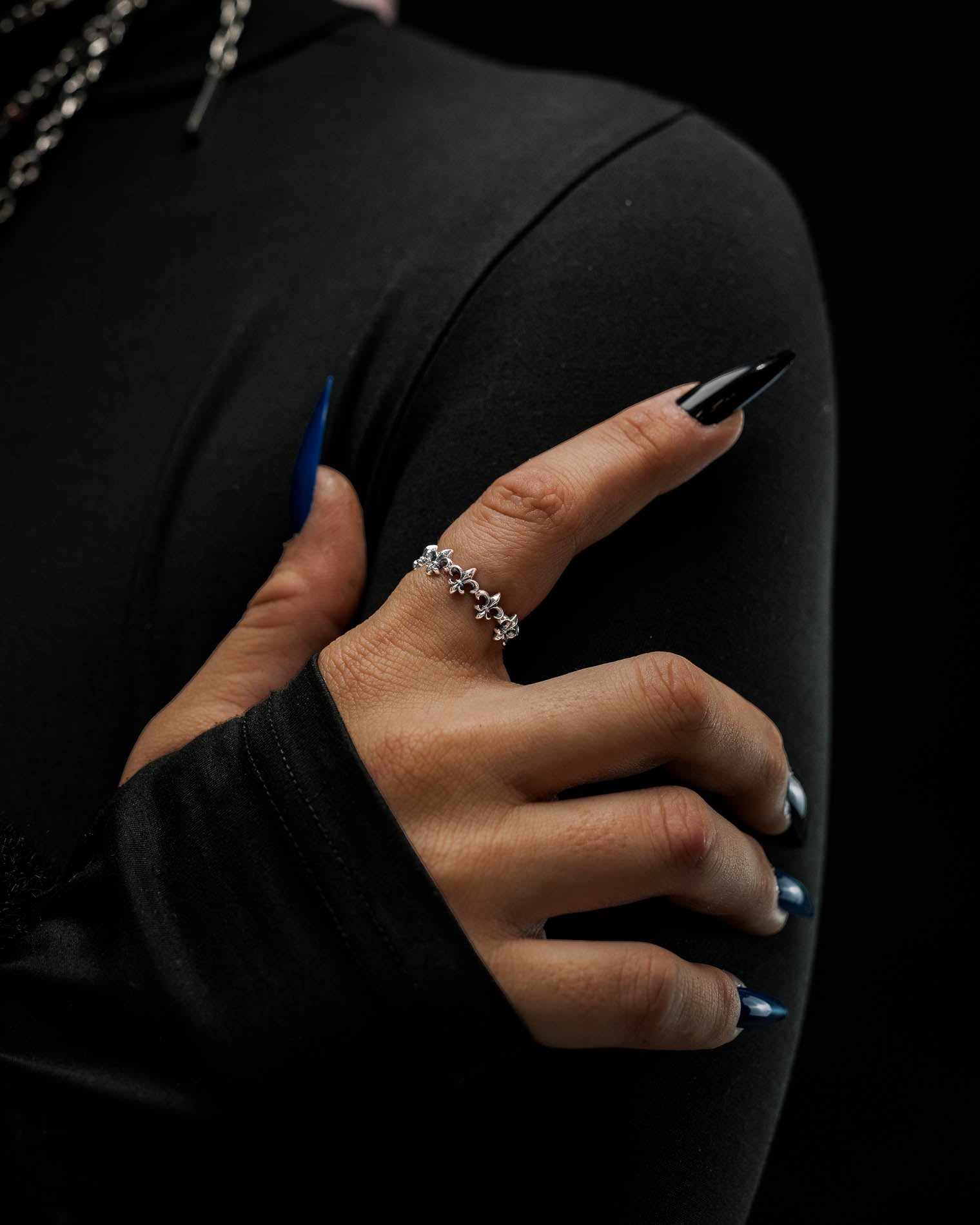 A hand with long, pointed black and blue nails wears the Statement Collective 925 Sterling Silver “Fleur De Lis” Ring, its antique silver finish adding a touch of elegance as the person, dressed in a fitted, long-sleeved black top, poses against a dark background.