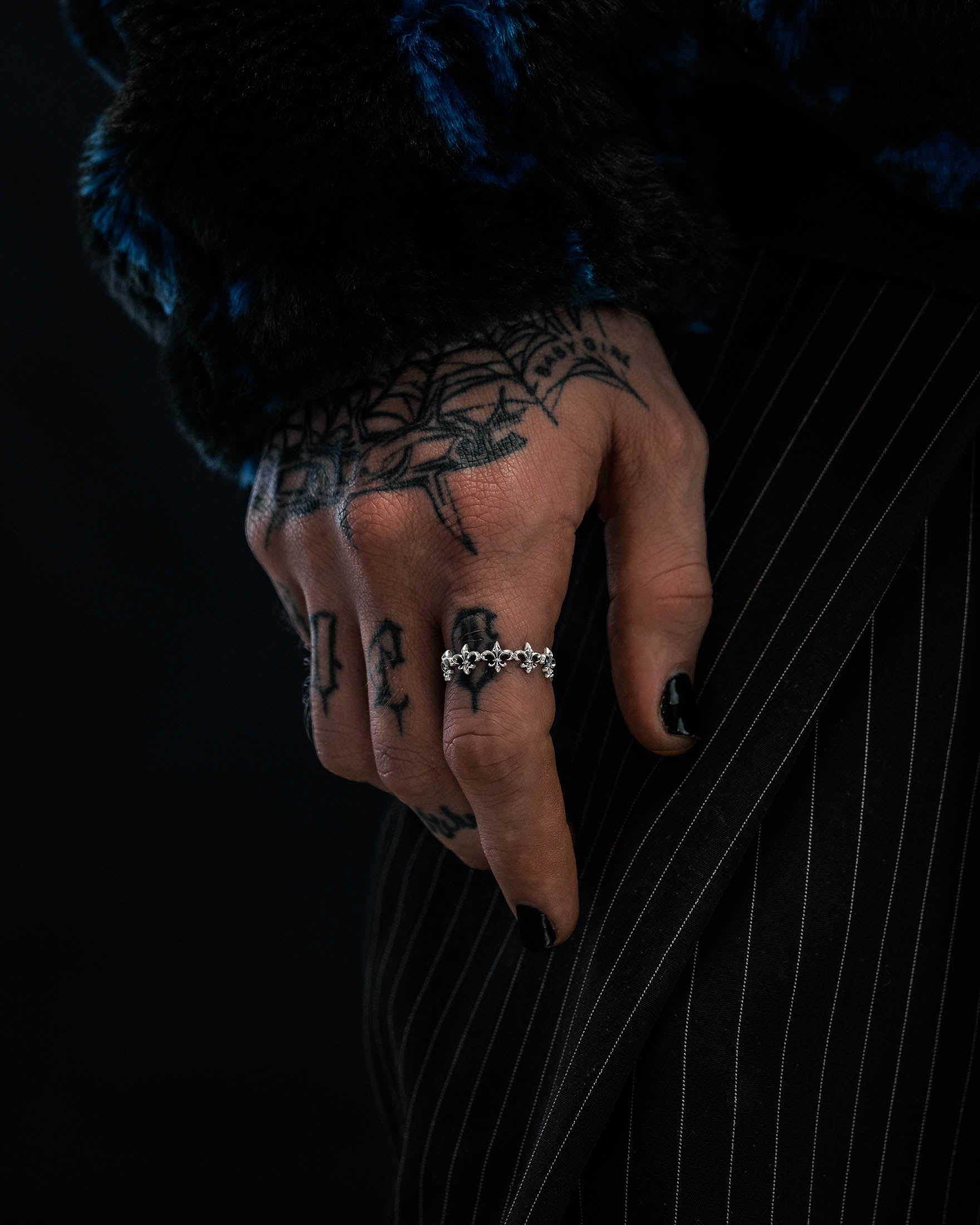 A tattooed hand with black nail polish wears the Statement Collective 925 Sterling Silver “Fleur De Lis” Ring, resting against pinstriped pants. The person is dressed in a dark, textured sleeve.