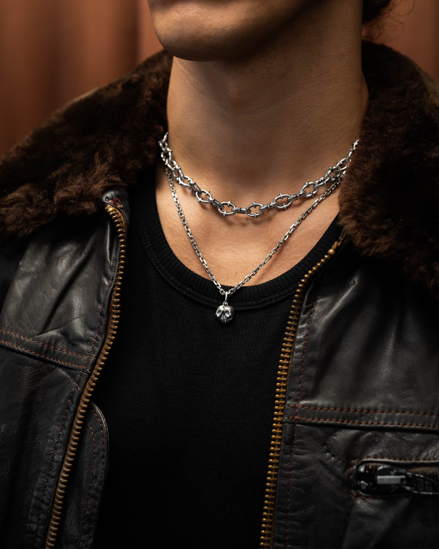 A person wears a brown leather jacket with a fur collar, a black shirt, and two silver necklaces—one is a chain link design and the other features the 925 Sterling Silver Skull Heart Pendant.