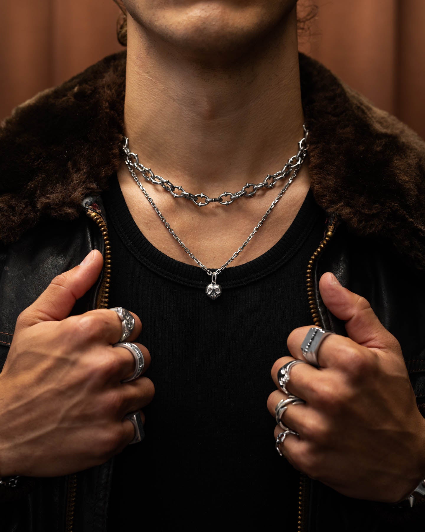 Wearing several sterling silver rings and the 925 Sterling Silver Skull Heart Pendant, a person grips the collar of a dark fur-lined jacket over a black shirt. The softly blurred brown background enhances their bold rock jewelry style.