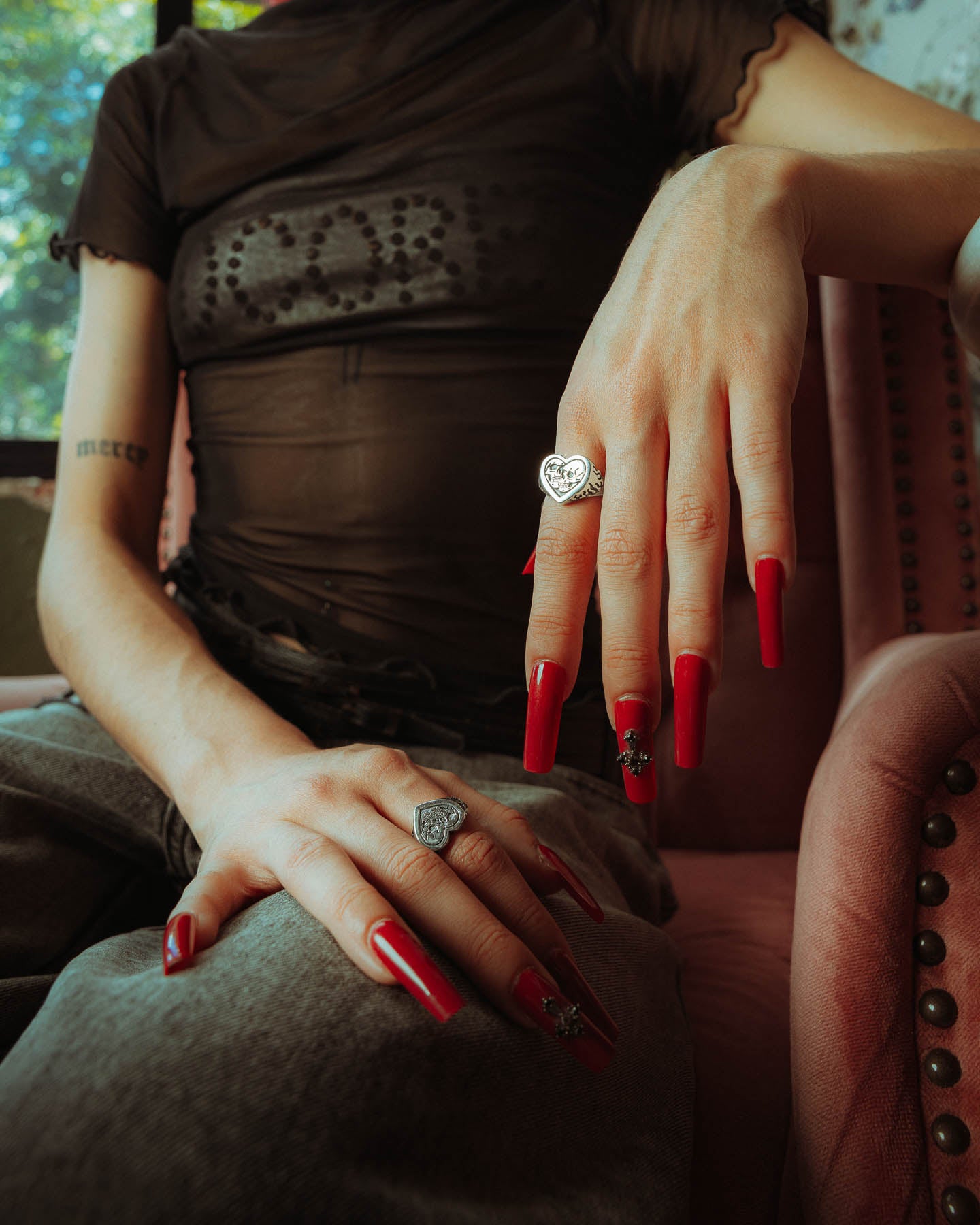 A person with long red nails, wearing a 925 Sterling Silver Skull Heart Ring, sits on a pink studded chair. They have tattoos on their arms and are dressed in a sheer black top and jeans.