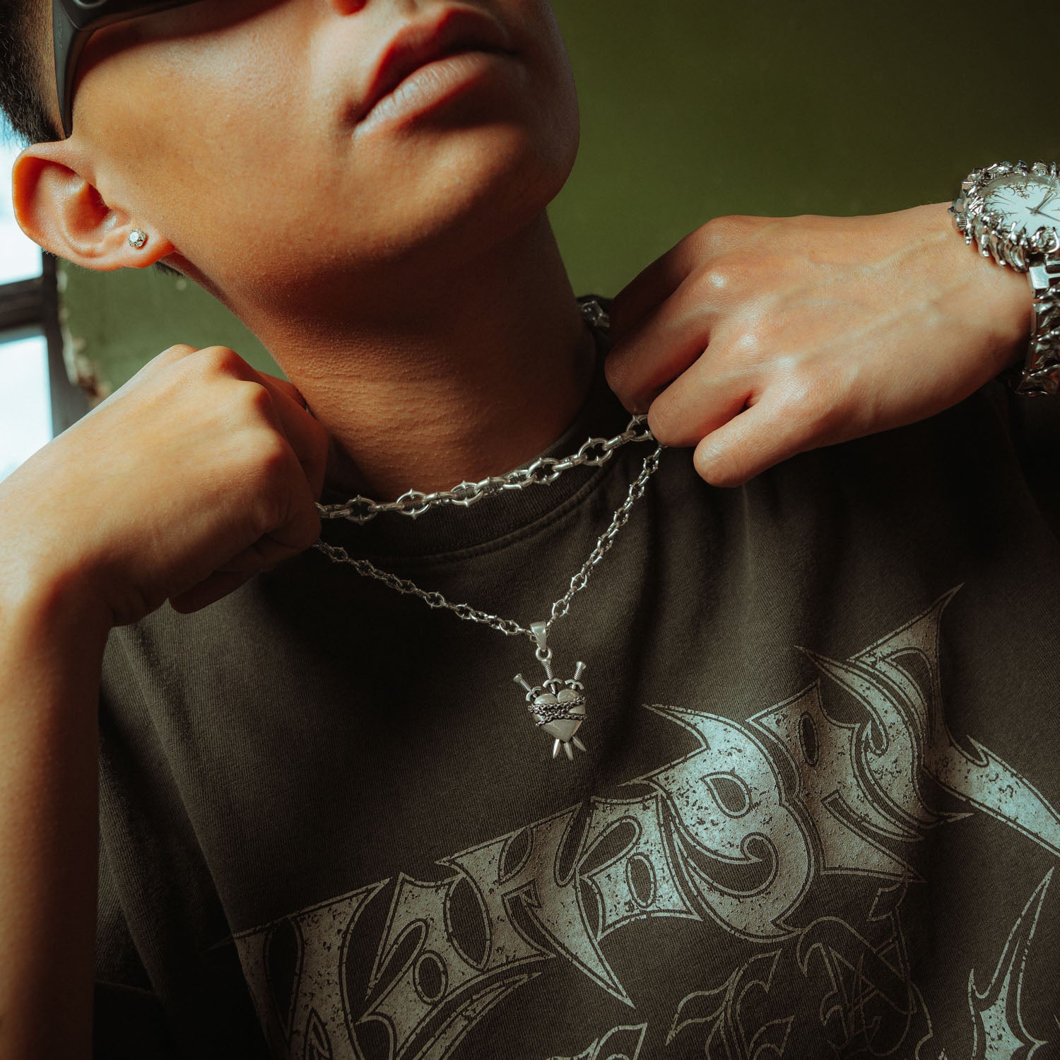 Silver heart pendant with three swords going through it and chain wrapped around it on a spiked chain with thicker spiked chain layered above on male model 