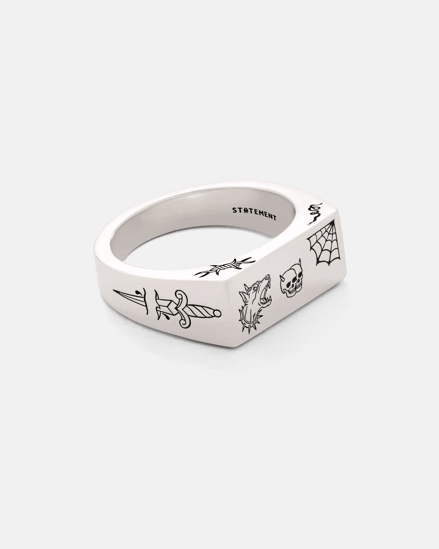 The 925 Sterling Silver Tattoo Signet Ring by Statement Collective features a flat top with black engraved dagger, wolf head, skull, and spider web designs. "STATEMENT" is engraved inside the band for a modern, edgy look.