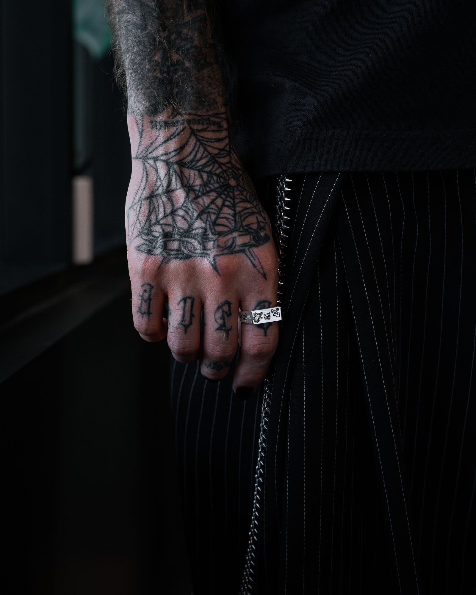 A tattooed hand with a spiderweb design and the word “PAST” on the fingers wears the Statement Collective 925 Sterling Silver Tattoo Signet Ring, paired with black pinstriped pants, a black shirt, and a side chain.