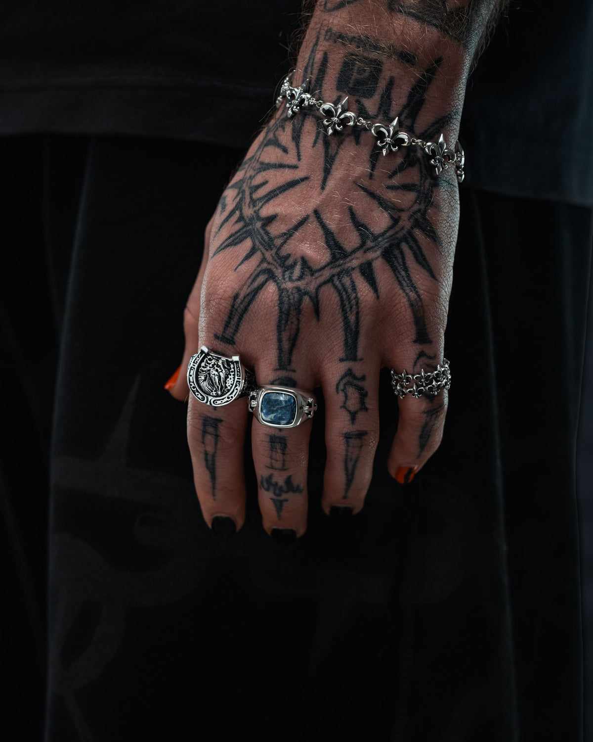 A tattooed hand with black painted nails showcases an array of rings, including the eye-catching 925 Sterling Silver Sodalite Stone Ring by Statement Collective, along with a bracelet. The bold, line-based tattoos cover the entire hand and fingers in a tattoo-inspired style. Set against a dark background, the focus remains on the hand adorned with striking sterling silver jewelry.