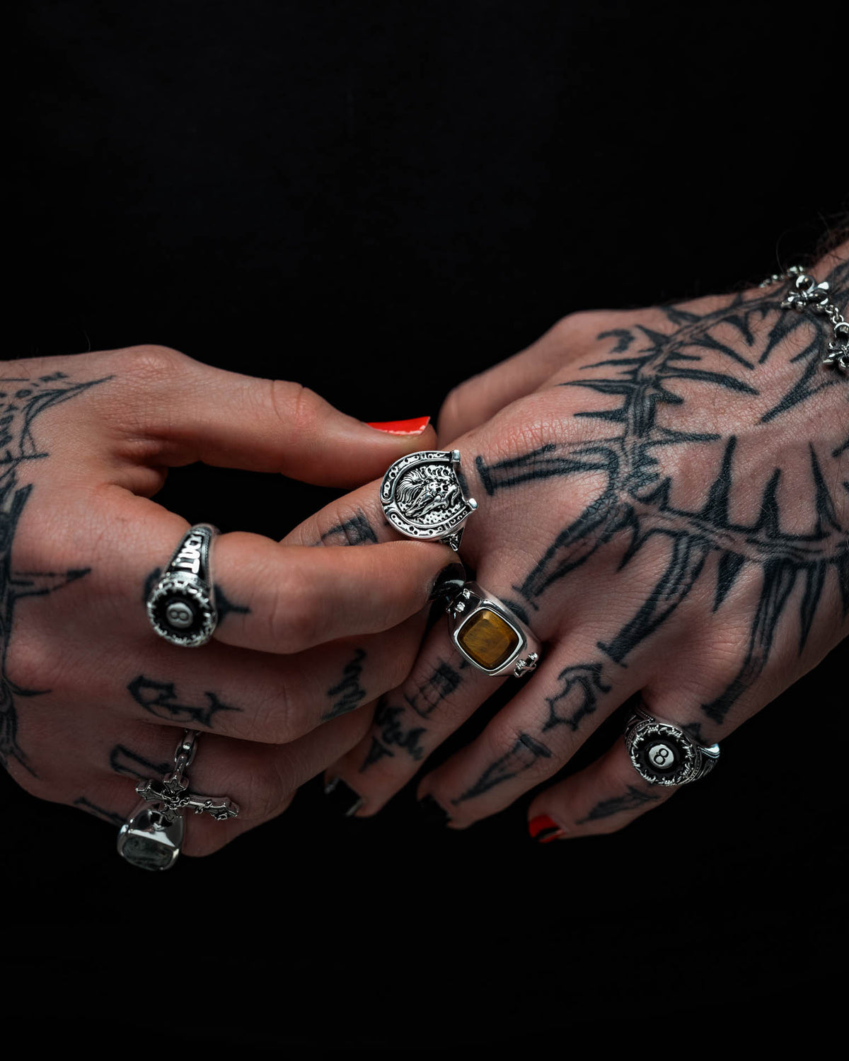 Hands tattooed and painted with black nail polish showcase multiple ornate rings by Statement Collective, including the 925 Sterling Silver Bronco Ring, along with other pieces of macabre jewellery and a bracelet, all set against a black background.