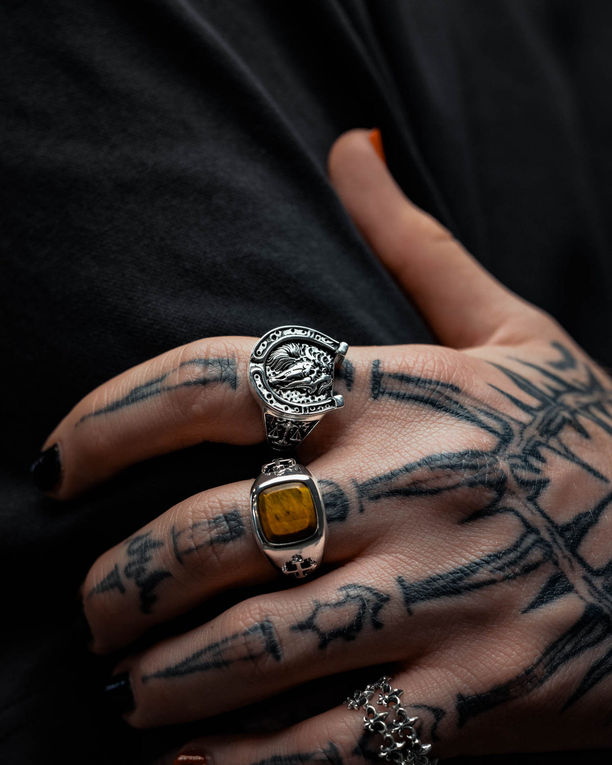 A tattooed hand with black nail polish wears two large silver rings, including the 925 Sterling Silver Bronco Ring by Statement Collective and another ring set with a yellow stone, showcasing striking macabre jewellery against a dark background.
