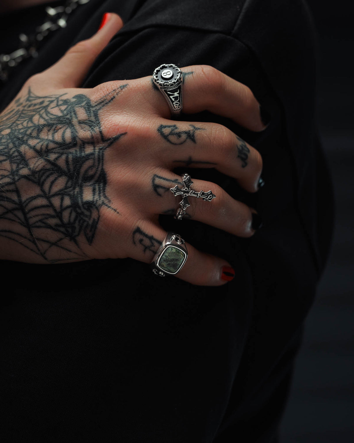 A tattooed hand with red nail polish rests on a black sleeve, showcasing several silver rings—including the Statement Collective 925 Sterling Silver Kambaba Jasper Stone Ring—crafted in sterling silver style. A spiderweb tattoo accentuates the edgy, tattoo-inspired aesthetic of these distinctive rings.