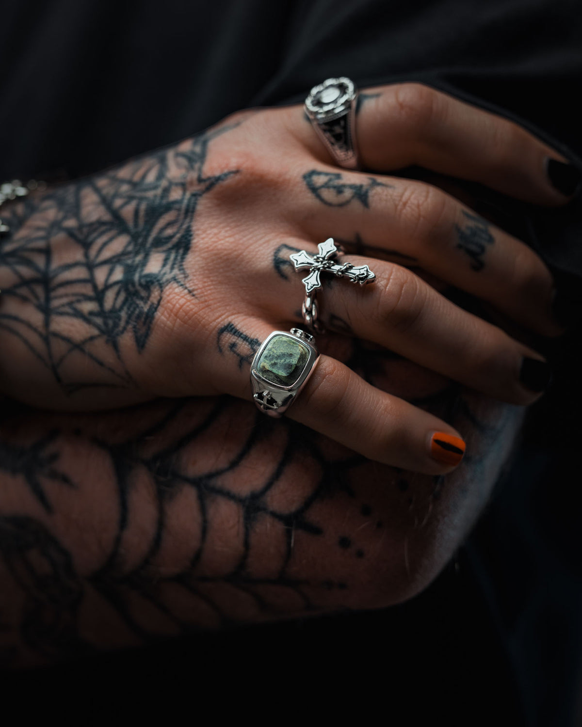 A tattooed hand with black nail polish and spiderweb tattoos showcases sterling silver jewelry, including a cross, a signet ring, and the Statement Collective 925 Sterling Silver Kambaba Jasper Stone Ring. One nail is painted orange. The hand rests on a black cloth.