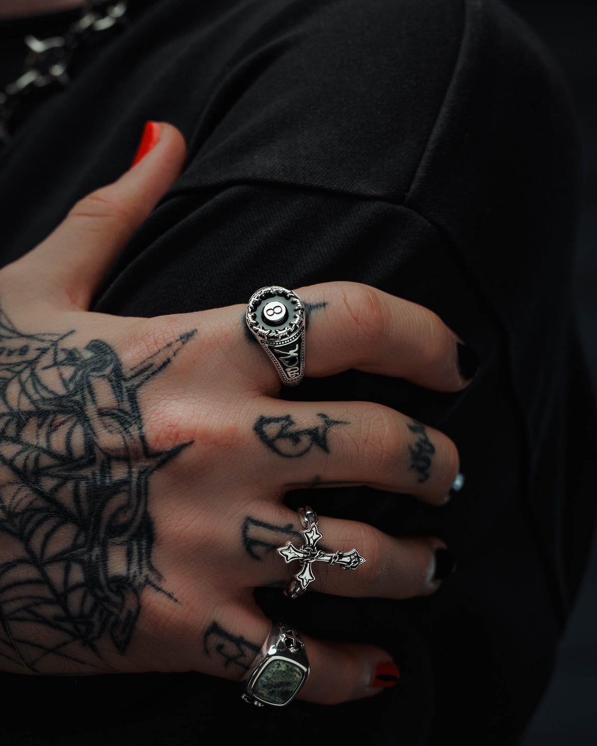 A hand adorned with spider web tattoos and black nail polish wears three silver rings, including the 925 Sterling Silver Lucky Shot Ring by Statement Collective—a striking piece featuring an 8-ball—alongside a cross ring and a square stone ring. Evoking tattoo-inspired jewelry, the hand grasps a piece of black fabric.