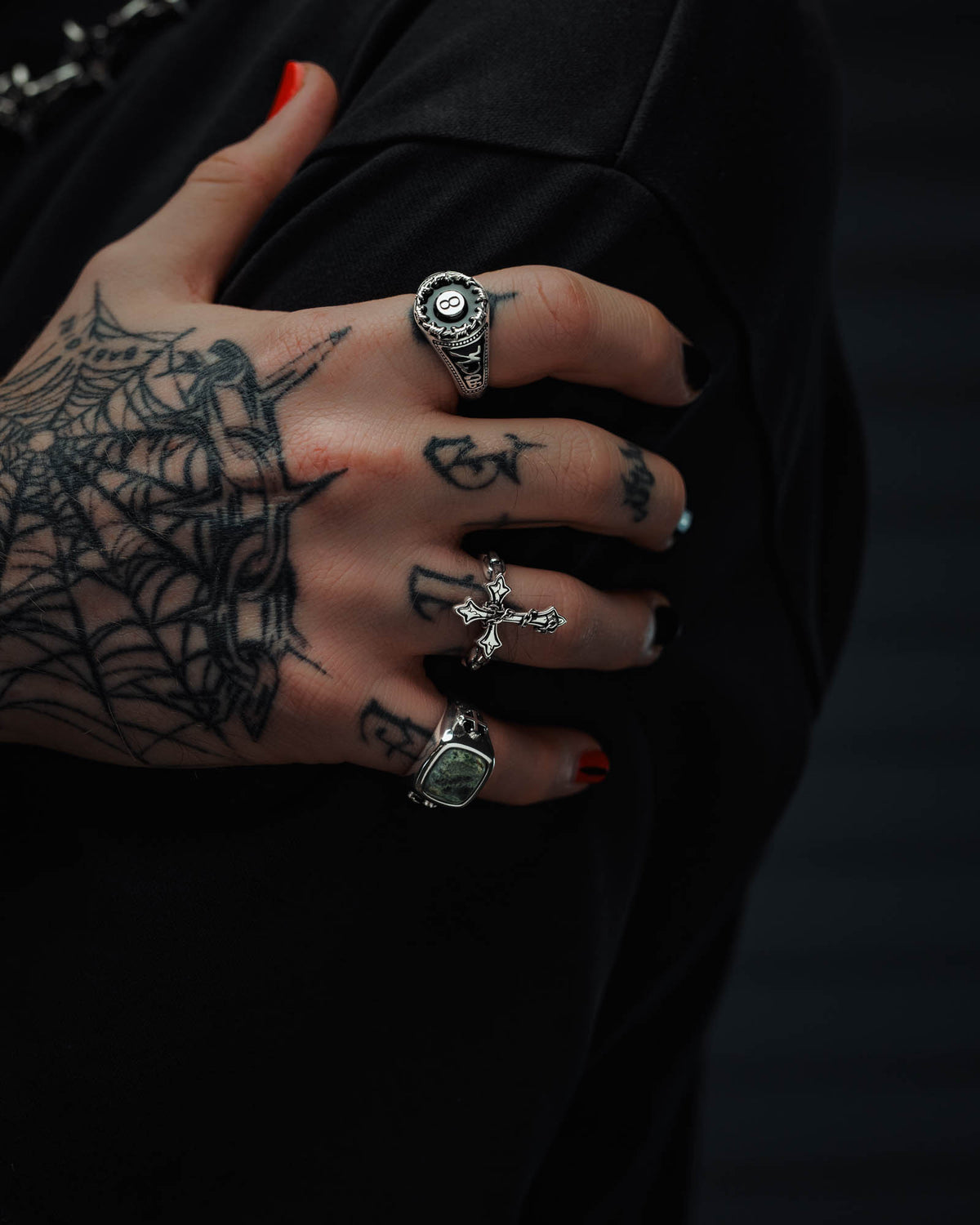 A hand with spiderweb tattoos showcases three silver rings against a dark background, including Statement Collective’s 925 Sterling Silver “The Ruins” Chained Cross Ring. The nails are painted black, with one red accent nail.