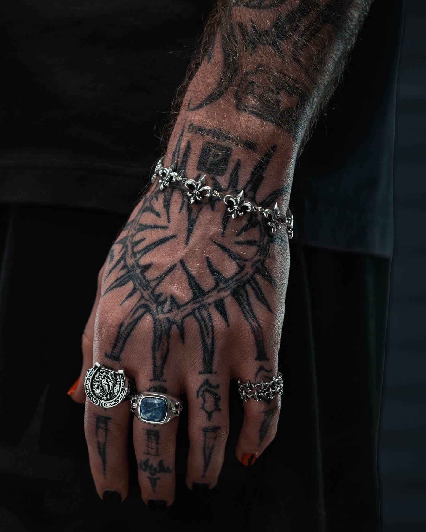A tattooed hand with black nail polish is adorned with several silver rings and the Statement Collective 925 Sterling Silver “Fleur De Lis” 15mm Bracelet, which features a distinct floral pattern. A prominent heart tattoo surrounded by rays and small human figures decorates the hand, set against a dark, blurred background.
