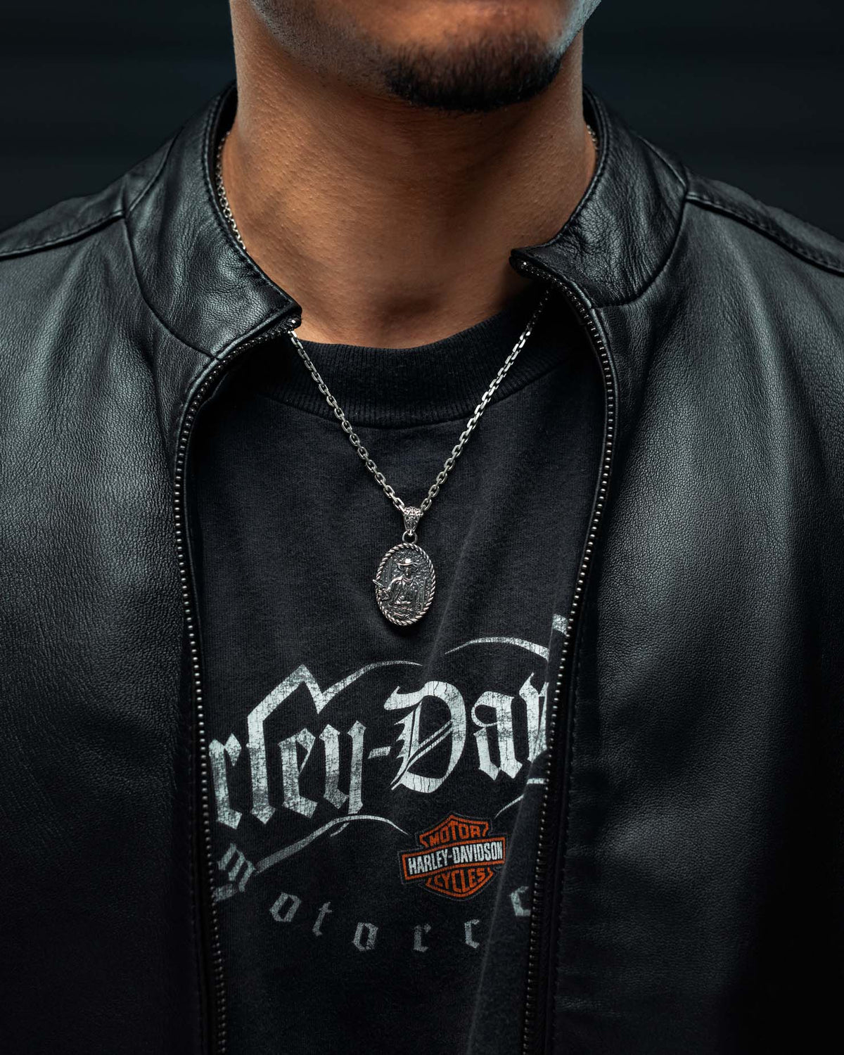 A person wearing a black leather jacket, a Harley-Davidson t-shirt, and the Statement Collective 925 Sterling Silver “Big Iron” Pendant. The image is cropped to show only the lower face, neck, and upper torso.