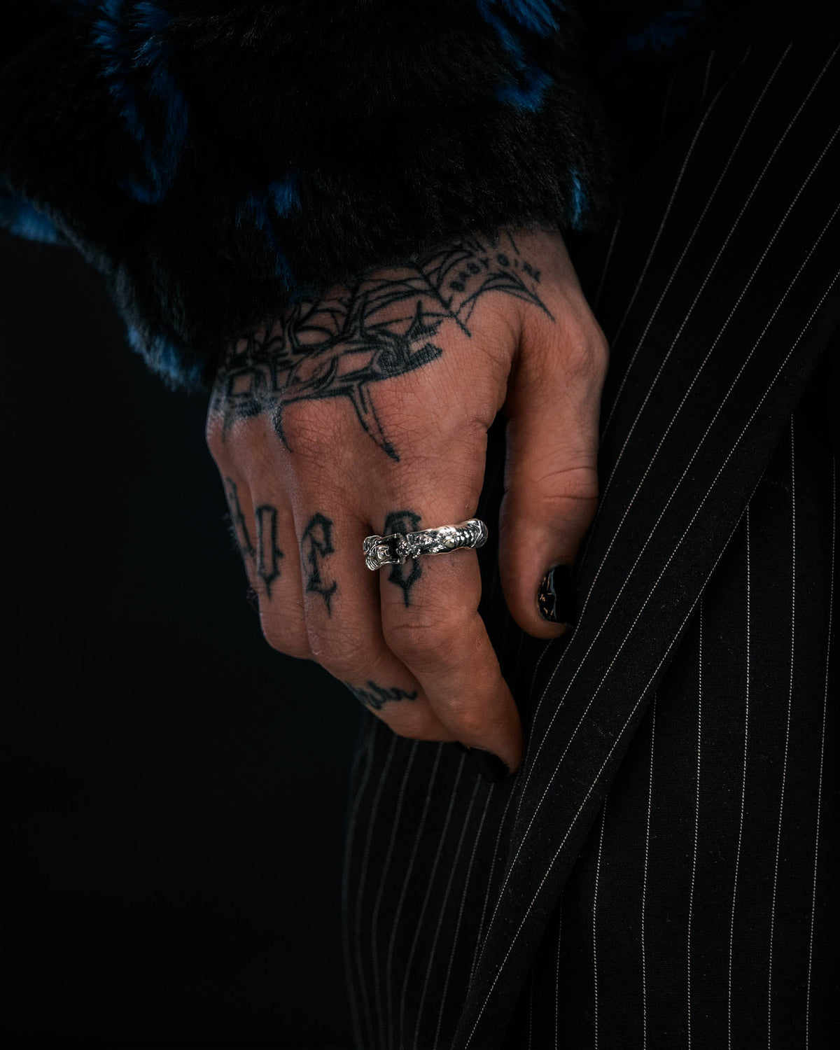 A tattooed hand with black-painted nails wears the Statement Collective 925 Sterling Silver Breaking Wheel Ring and rests on pinstriped pants. The person is also wearing a dark, textured jacket.