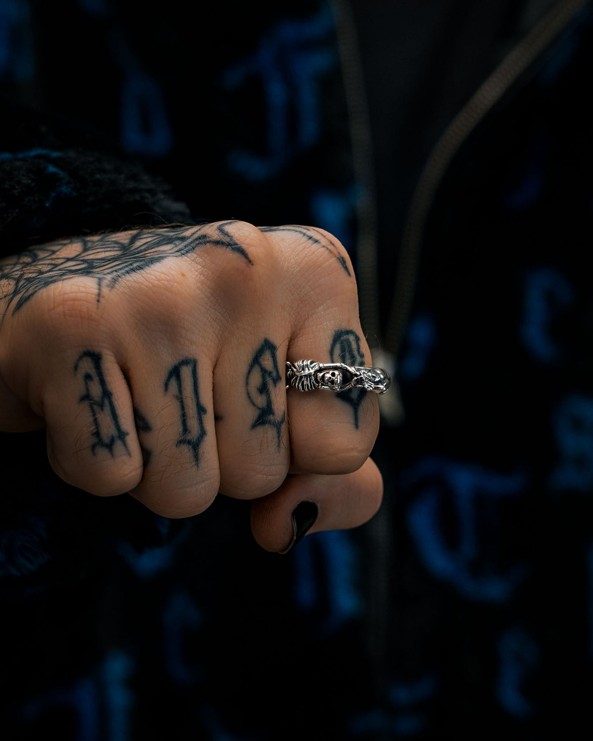 A close-up of a fist with “L I F E” tattooed on the fingers, wearing Statement Collective’s 925 Sterling Silver Breaking Wheel Ring on the middle finger and displaying a web tattoo on the hand. The person is dressed in a black and blue patterned jacket.