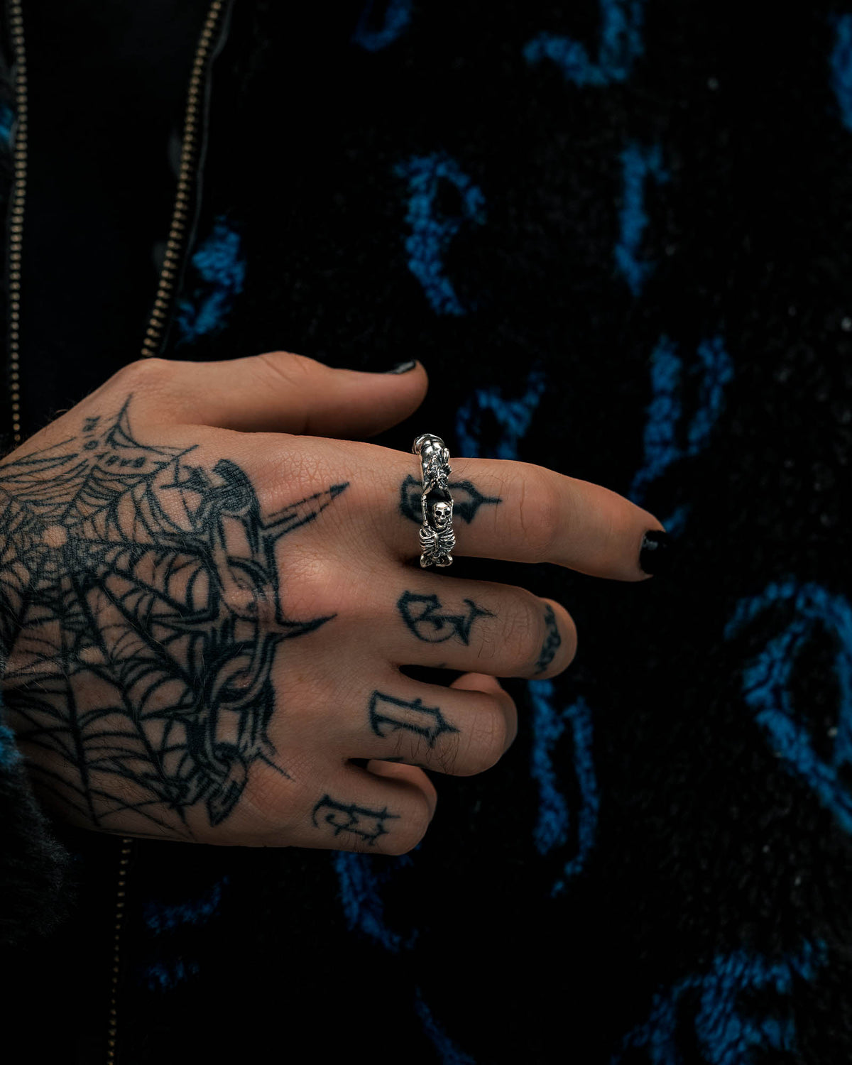 A tattooed hand with a spider web design wears the Statement Collective 925 Sterling Silver Breaking Wheel Ring on the middle finger, set against a dark background with blue patterns.