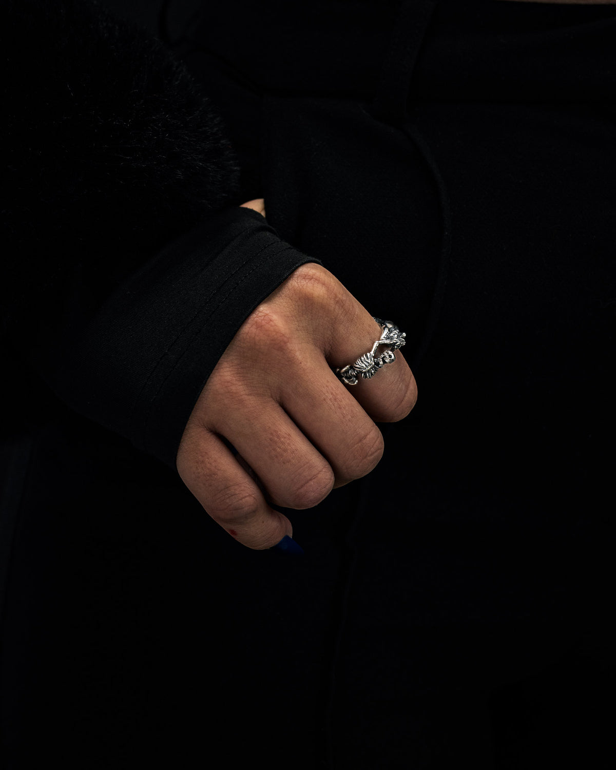 A close-up of a hand wearing the Statement Collective 925 Sterling Silver Breaking Wheel Ring, set against a black outfit. The thumb is painted with blue nail polish, and the sleeve features a furry black cuff.