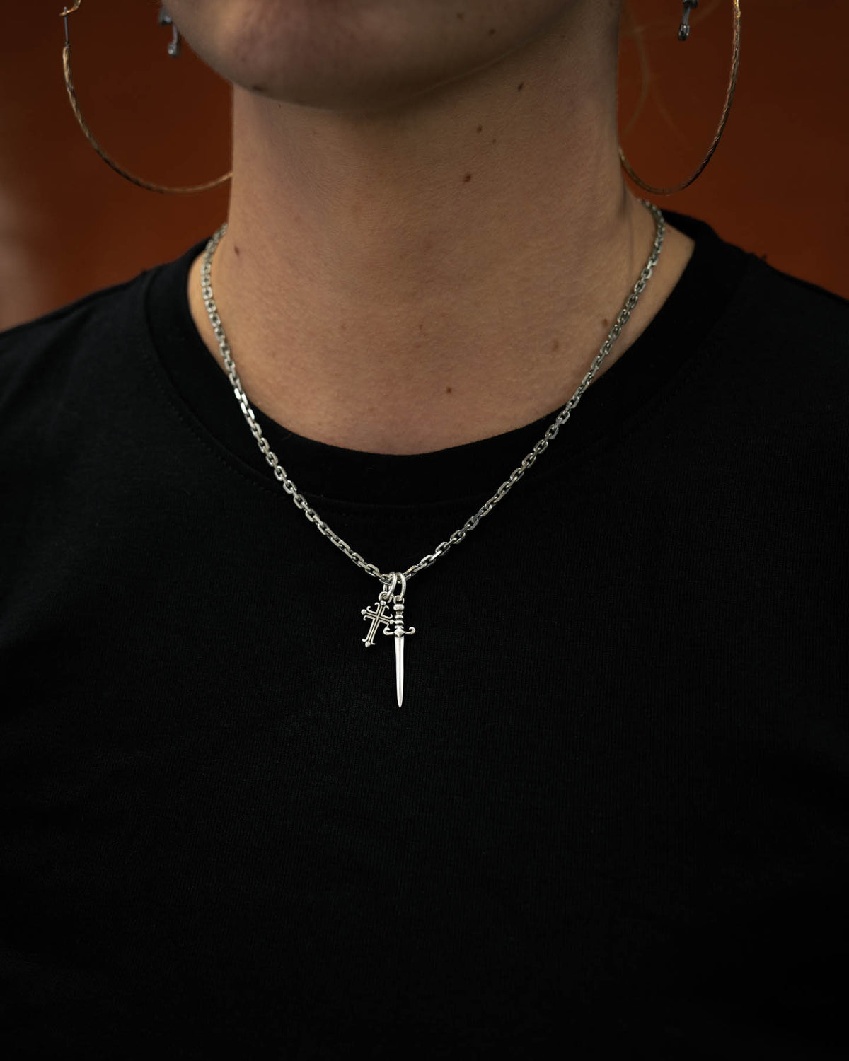 A person shown from the shoulders up, dressed in a plain black shirt, wears large hoop earrings and the Statement Collective 925 Sterling Silver Cross & Dagger Necklace, adding a rock jewelry vibe with its striking sword and cross charms.