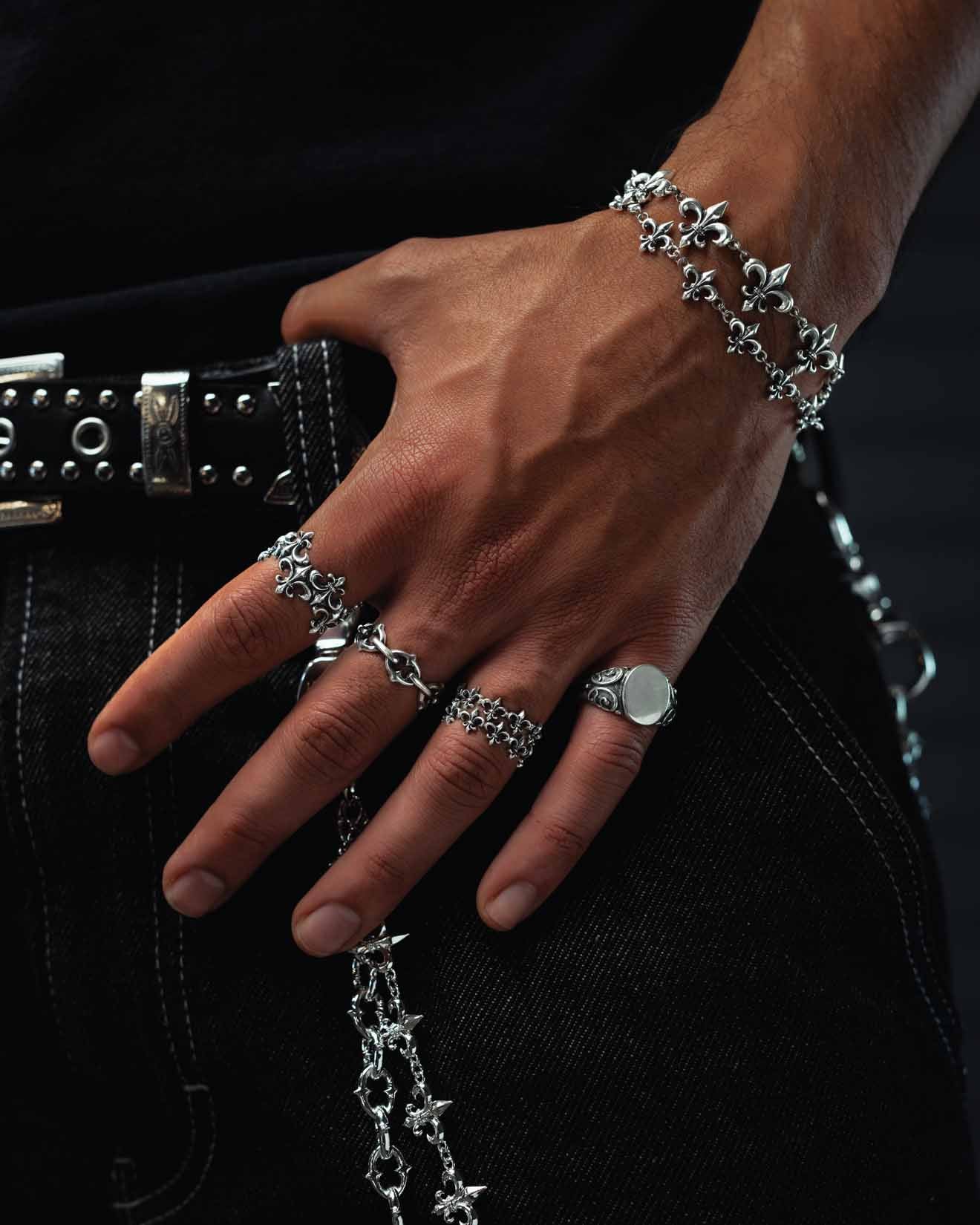 A close-up of a hand adorned with multiple silver rings—including the Statement Collective 925 Sterling Silver “Fleur De Lis” Ring—paired with matching bracelets, all showcasing detailed fleur-de-lis designs. The accessories stand out against black jeans with a studded belt and chain.