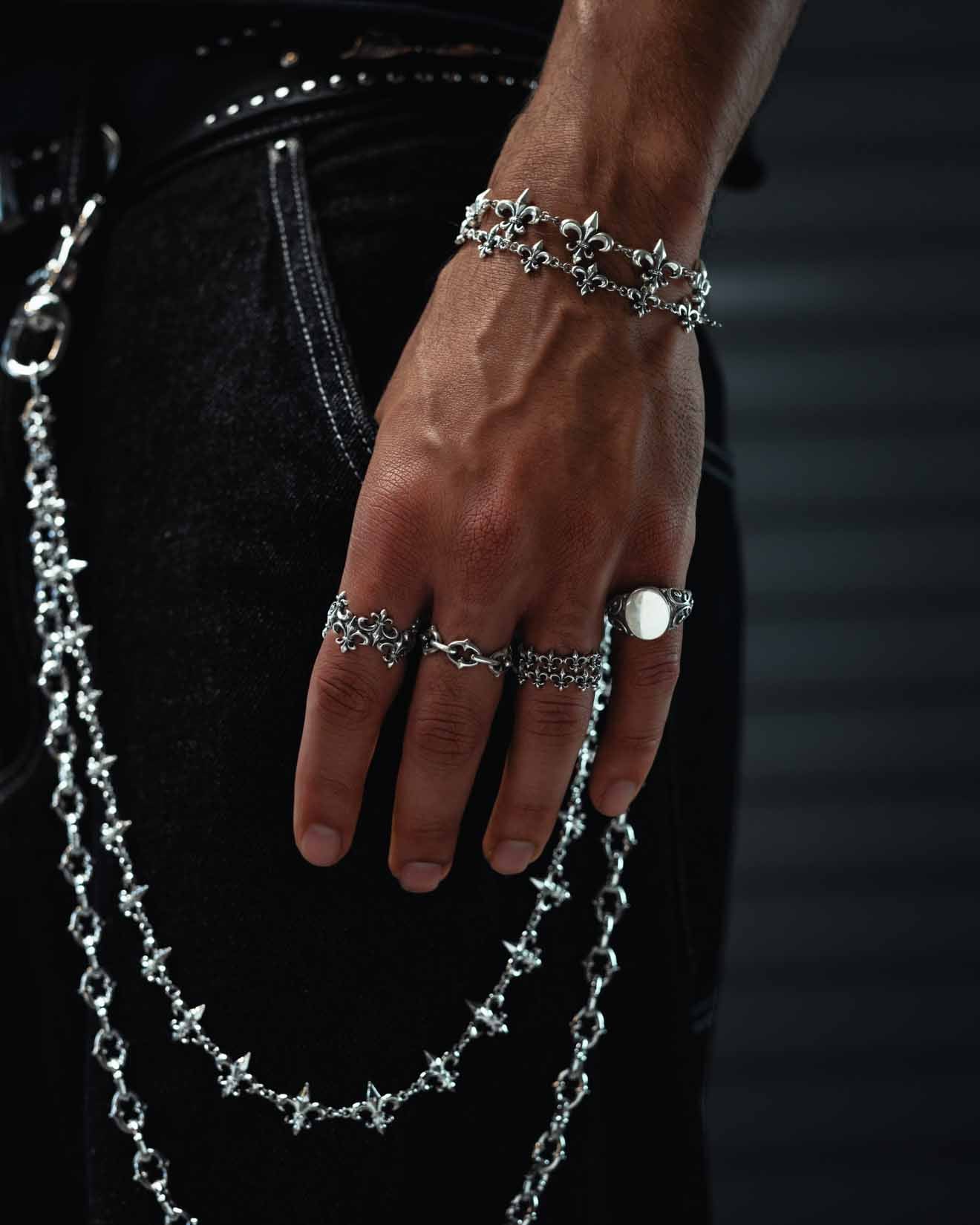 A close-up of a hand adorned with vintage silver jewelry, featuring the 925 Sterling Silver “Crested” Fleur De Lis Ring by Statement Collective, paired with matching bracelets and resting on dark jeans accented with silver chains at the waistband.
