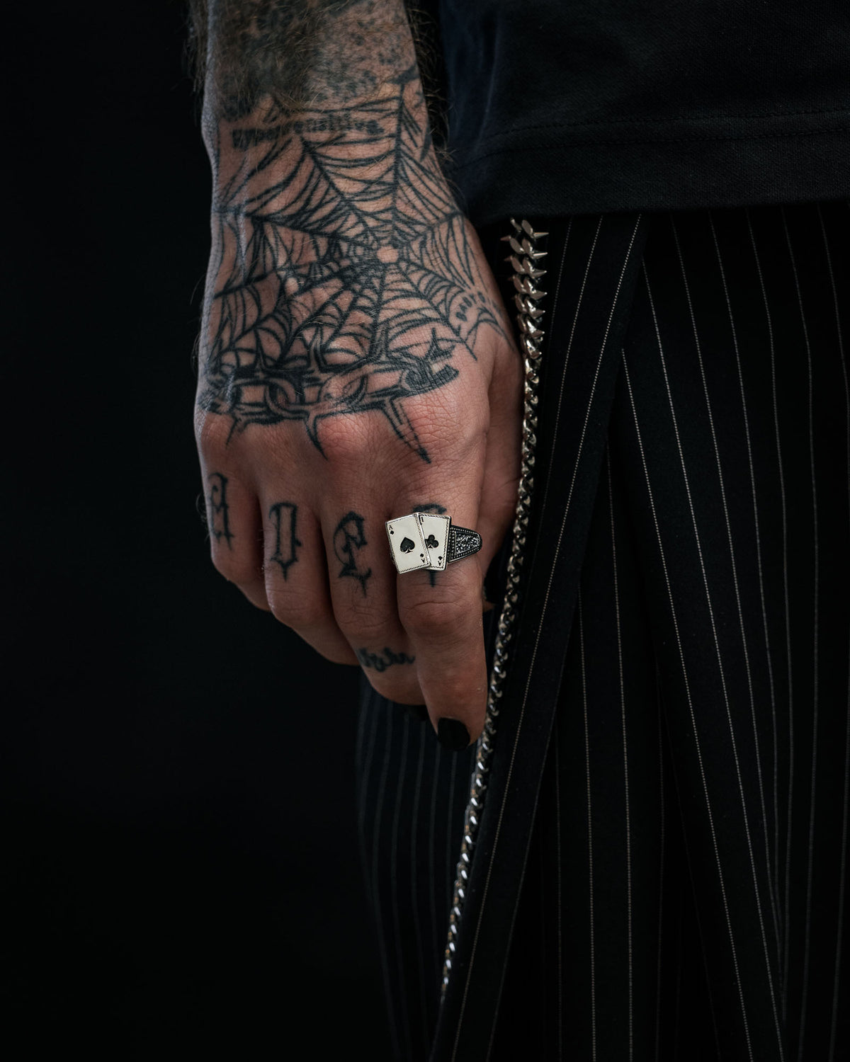 A tattooed hand with a spiderweb design showcases the Statement Collective 925 Sterling Silver Pocket Aces Ring, resting against pinstriped pants with a visible chain for an edgy, standout jewellery statement.