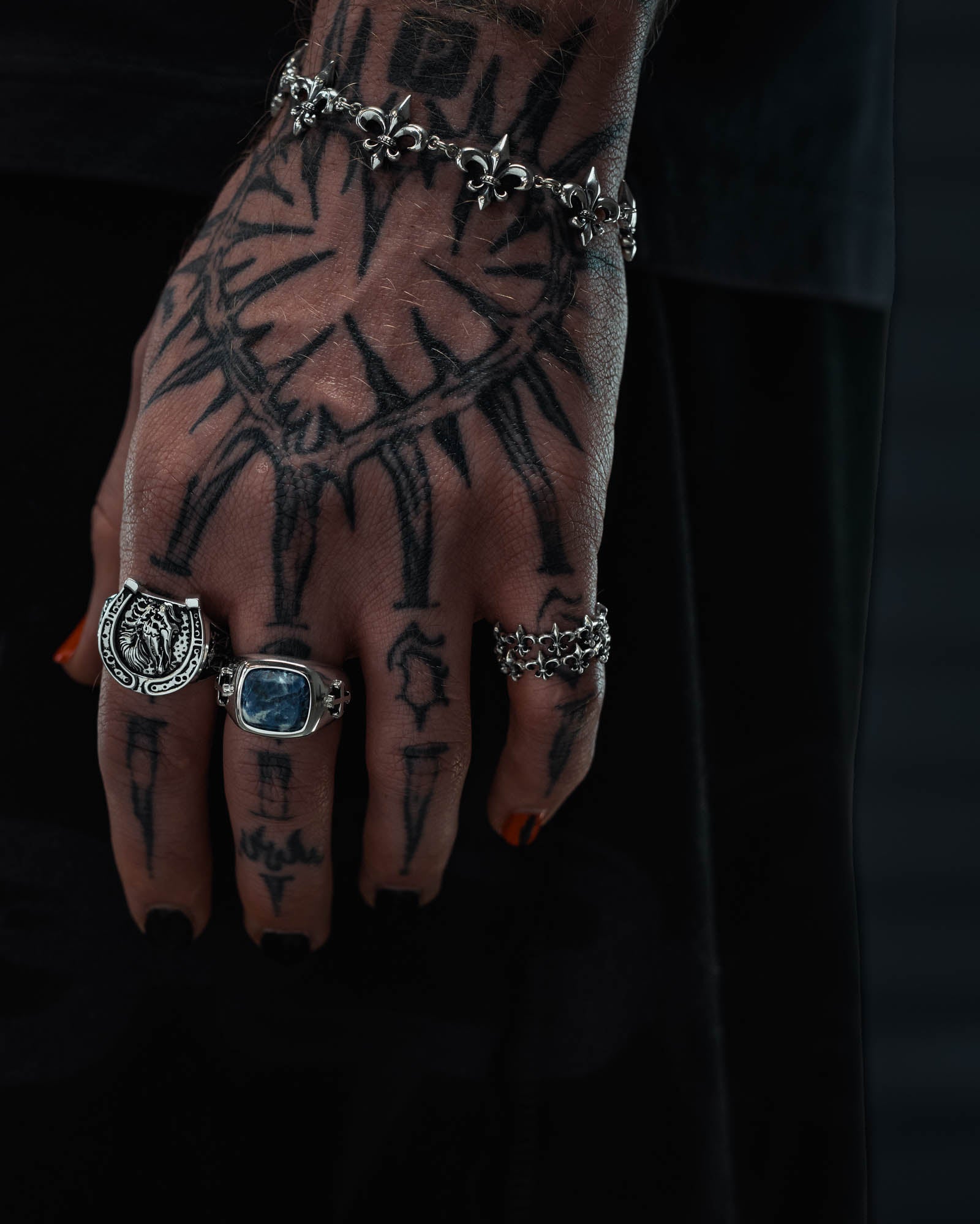 A tattooed hand, its fingernails painted black, is adorned with the Statement Collective 925 Sterling Silver “Fleur De Lis” Ring and a bracelet. The tattoos form a clock-like pattern, while the dark background accentuates the intricate fleur de lis details of the antique-inspired silver ring.