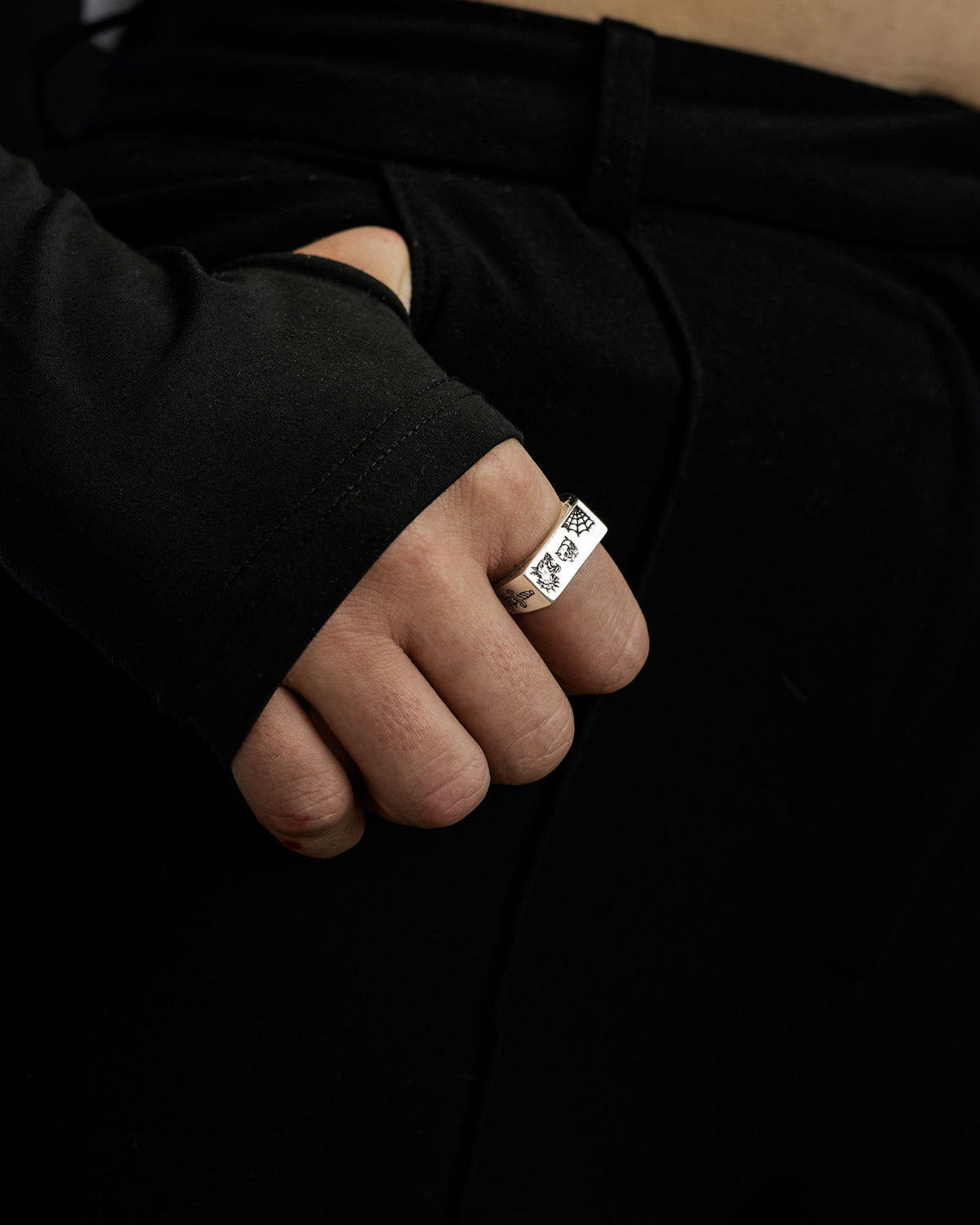 A hand adorned with the Statement Collective 925 Sterling Silver Tattoo Signet Ring rests on the waistband of black pants. The person wears a black long-sleeve top with a thumbhole, exposing only the fingers and part of the palm.