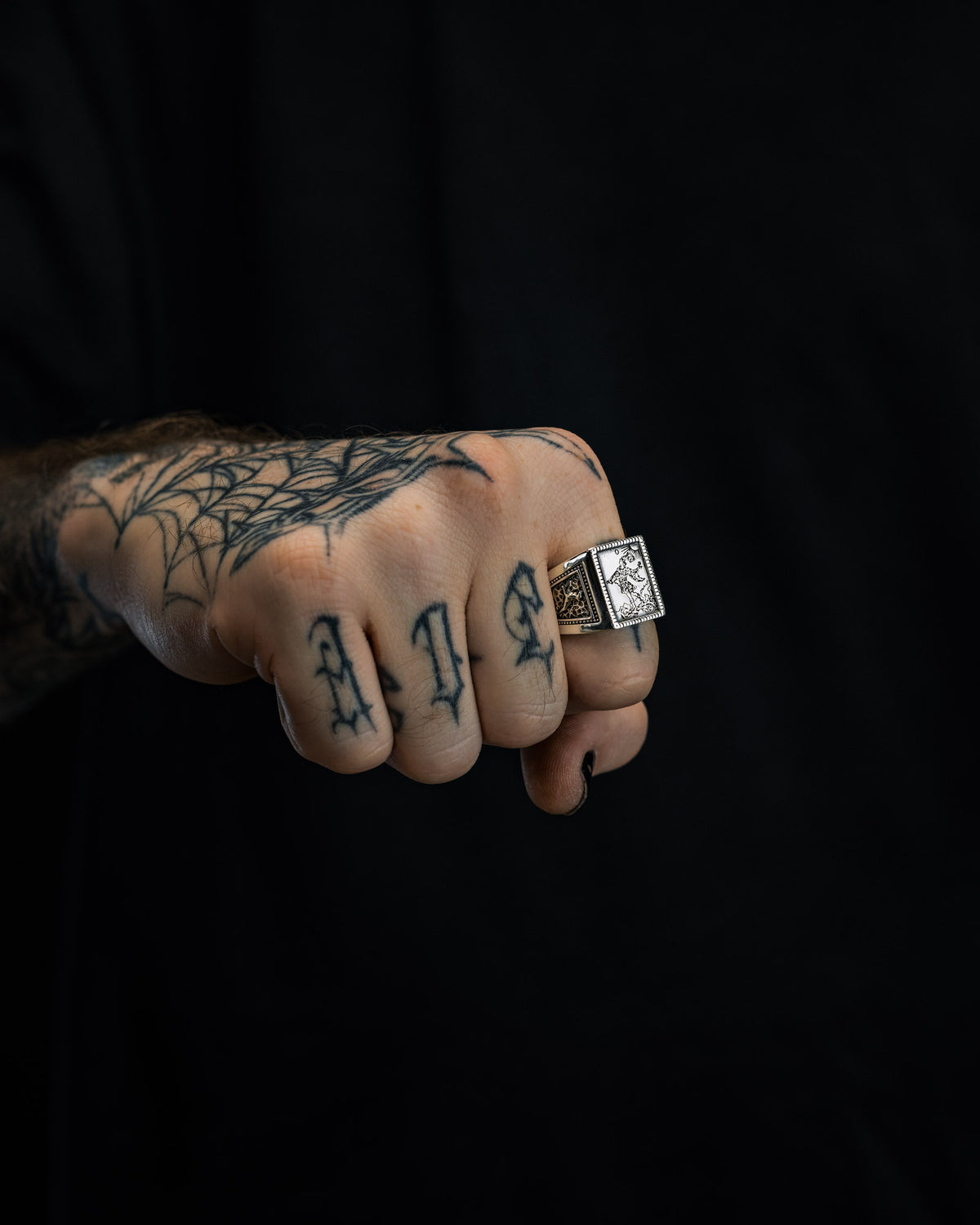 A tattooed hand with a spiderweb on the wrist and “ALS” on the fingers, wearing the Statement Collective 925 Sterling Silver “Try Hard Fool” Ring, is shown against a black background.